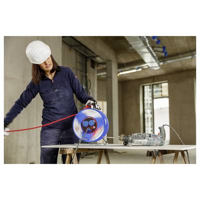 Bauarbeiter in Schutzhelm, der eine blaue Kabelrolle neben einem Bohrer auf Böcken an einer unfertigen Innenbaustelle handhabt.