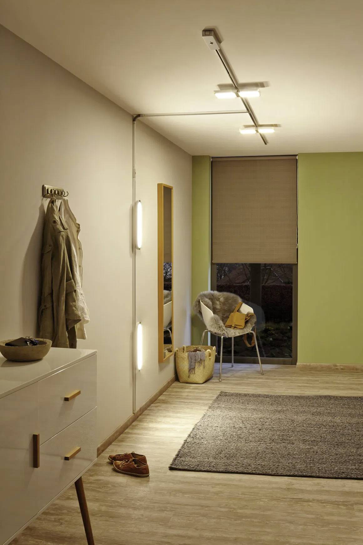 Ein Wohnzimmer mit einer beigen Wand, an der ein Mantel hängt. Ein Stuhl mit Decke, Spiegel, Lampe auf Kommode und ein Teppich auf Holzboden.