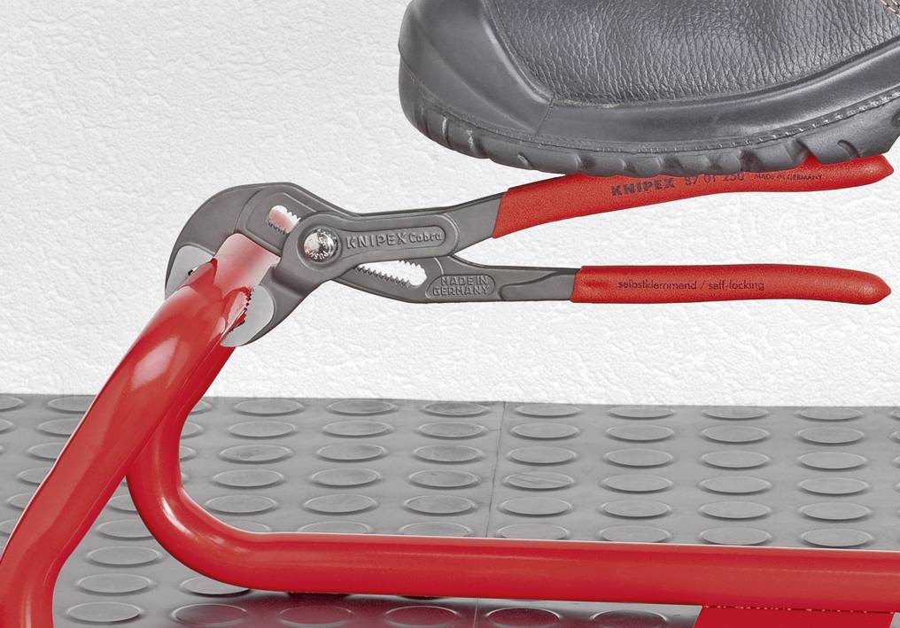 Eine rote Greifzange hält selbsttätig an einem runden Metallrohr fest. Ein schwarzer Schuh drückt auf den Zangenmechanismus.