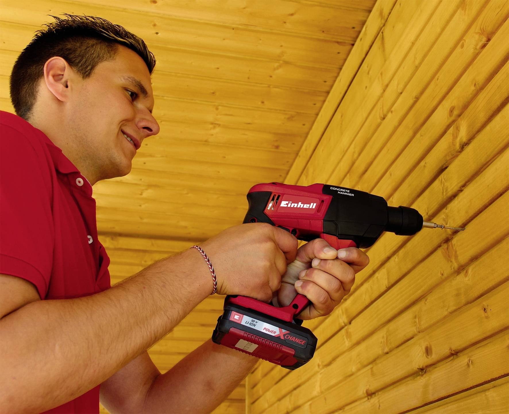 Ein Mann bohrt mit einer roten Bohrmaschine in eine Holzwand. Er trägt ein rotes Hemd und scheint konzentriert zu arbeiten.