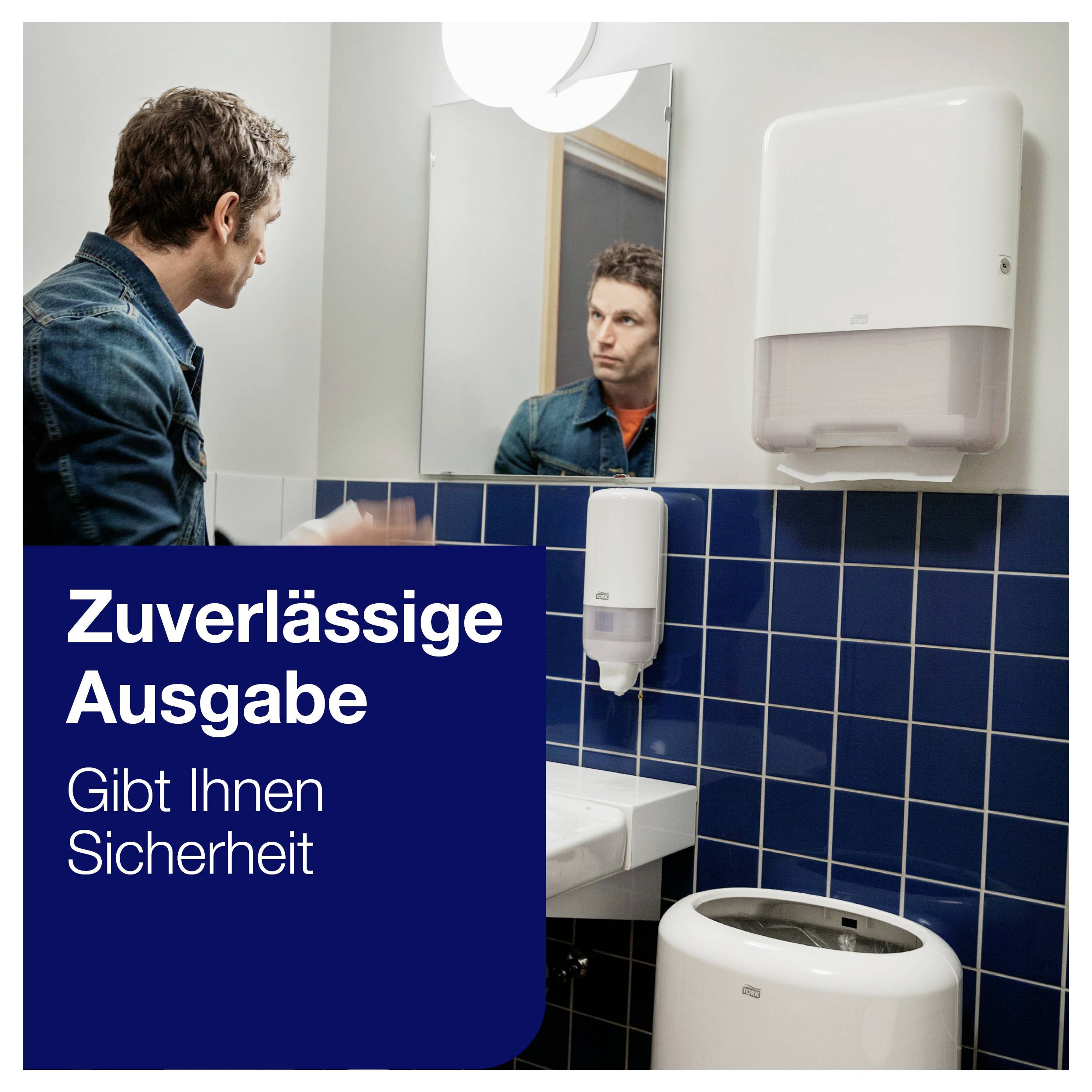 Ein Mann steht in einem Badezimmer vor einem Spiegel. Im Vordergrund ist ein Papierhandtuchspender und ein Waschbecken zu sehen. Text: "Zuverlässige Ausgabe - Gibt Ihnen Sicherheit".
