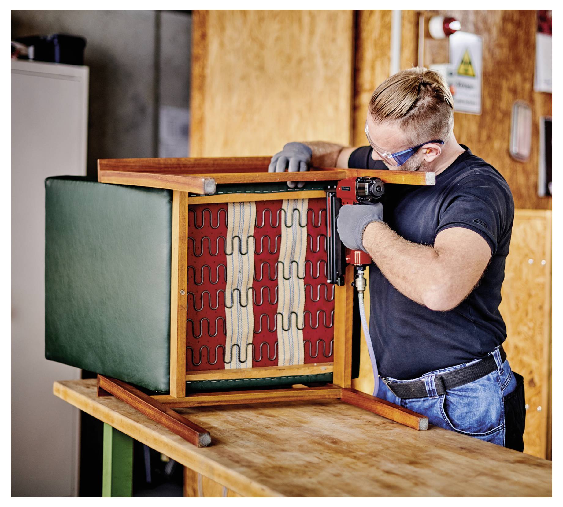 Ein Mann, der Schutzbrille und Handschuhe trägt, befestigt ein Holzgestell eines Stuhls und zeigt dabei die Sitzfedern und die Polsterung während der Möbelreparatur.