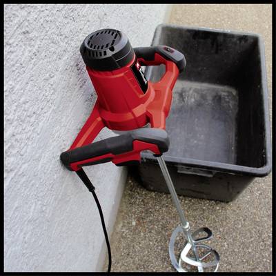 'Ein roter elektrischer Betonmischer mit Rührstab lehnt an einer Wand neben einer schwarzen Plastikwanne auf einem Betonboden.'