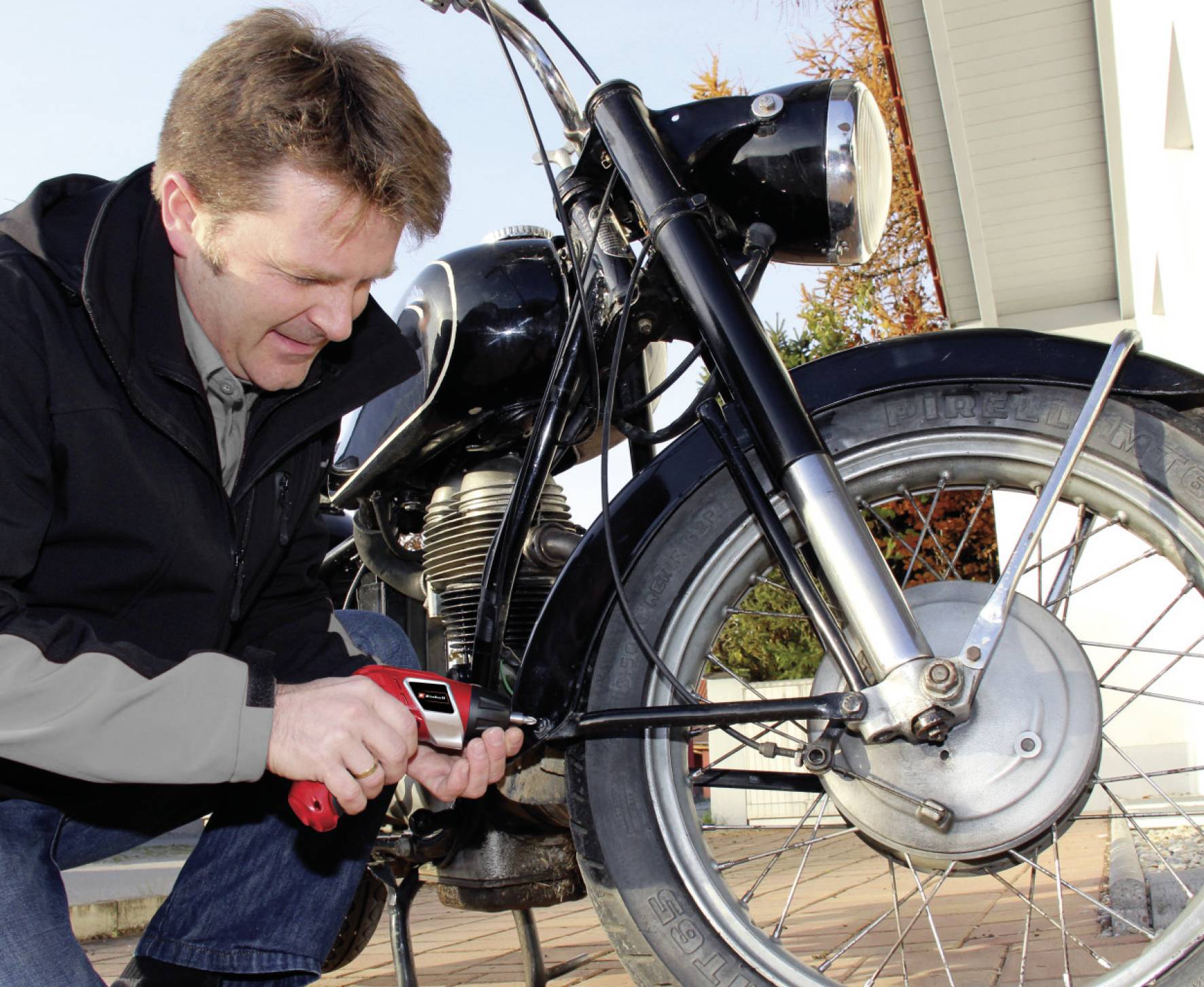 Ein Mann repariert ein Motorrad im Freien. Er benutzt ein Werkzeug zur Befestigung von Teilen am Vorderrad. Es ist sonnig.