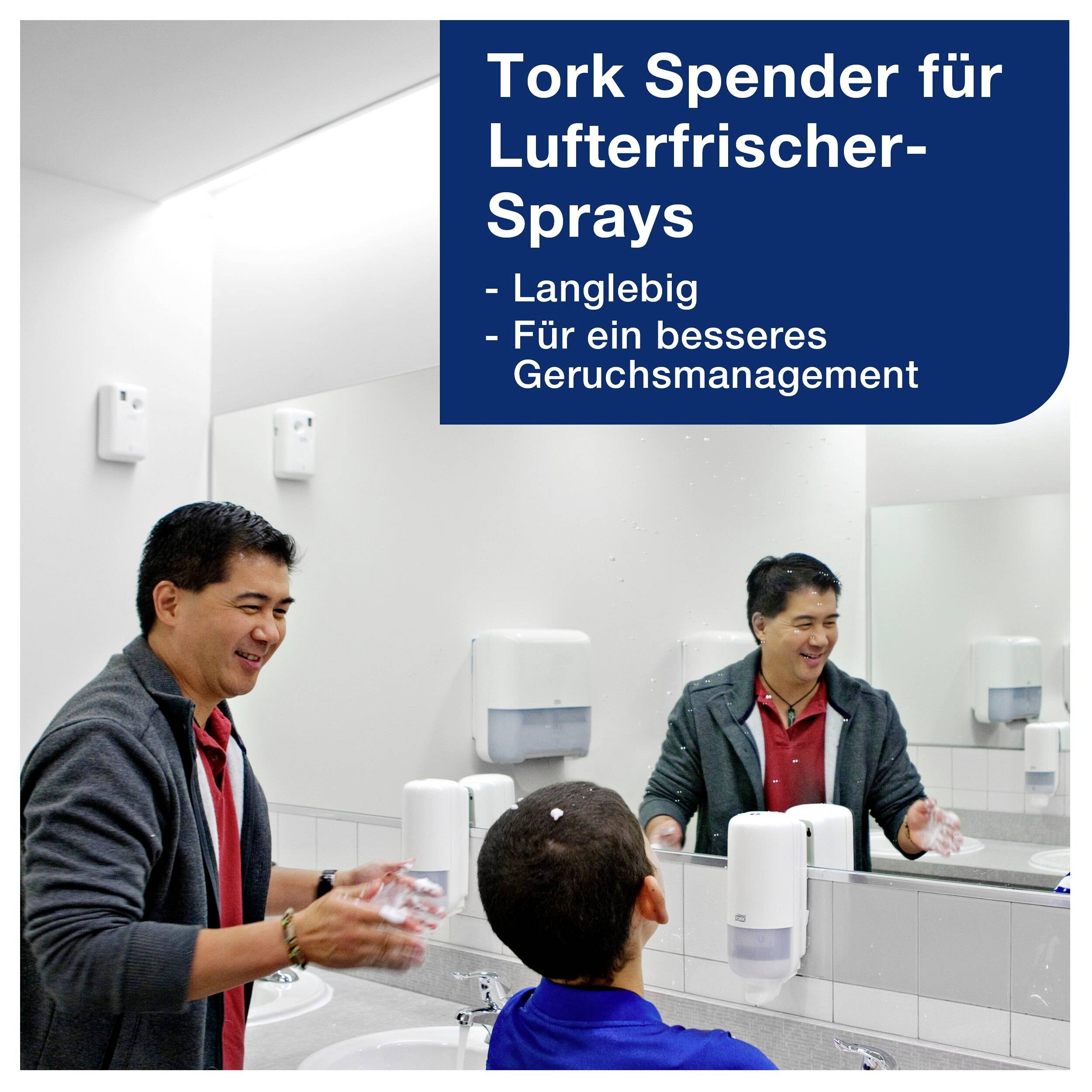 Eine Person wäscht sich die Hände in einem Badezimmer. An der Wand hängt ein Luftsprayspender mit der Aufschrift: 'Tork Spender für Lufterfrischer-Sprays - Langlebig - Für ein besseres Geruchsmanagement'.