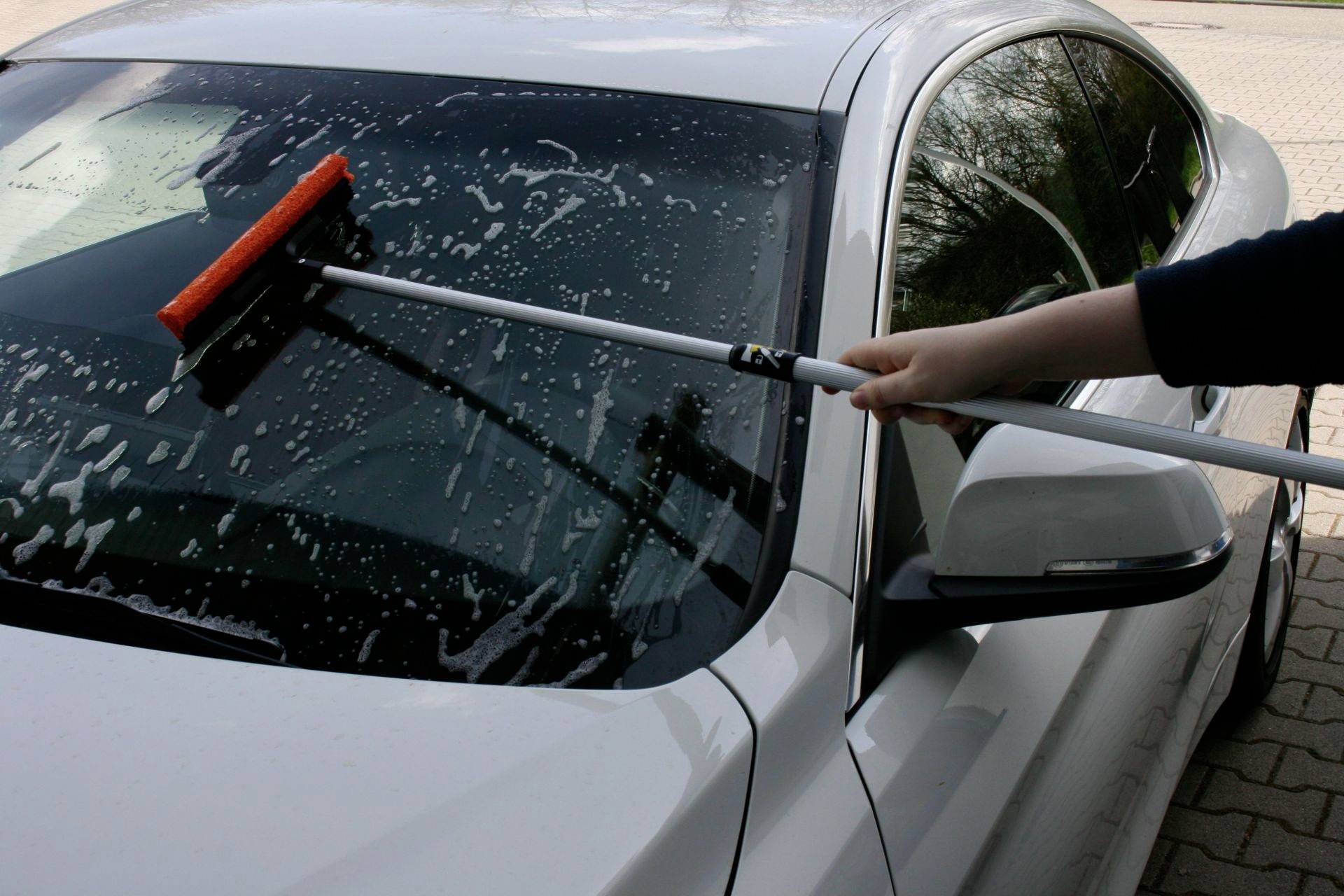 Eine Person reinigt die Windschutzscheibe eines weißen Autos mit einem langen Besenstiel und Schaum.
