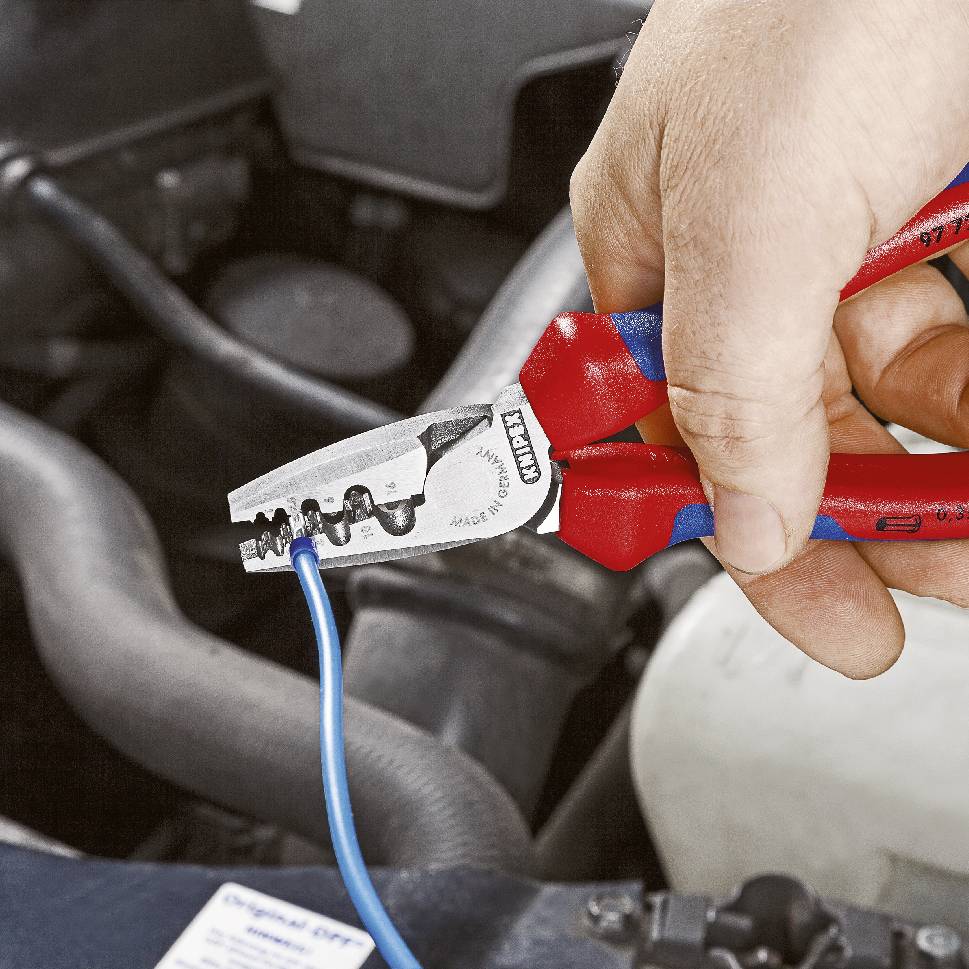 Person benutzt rote Drahtschneider, um ein blaues Kabel in einem Fahrzeugmotorraum zu schneiden.