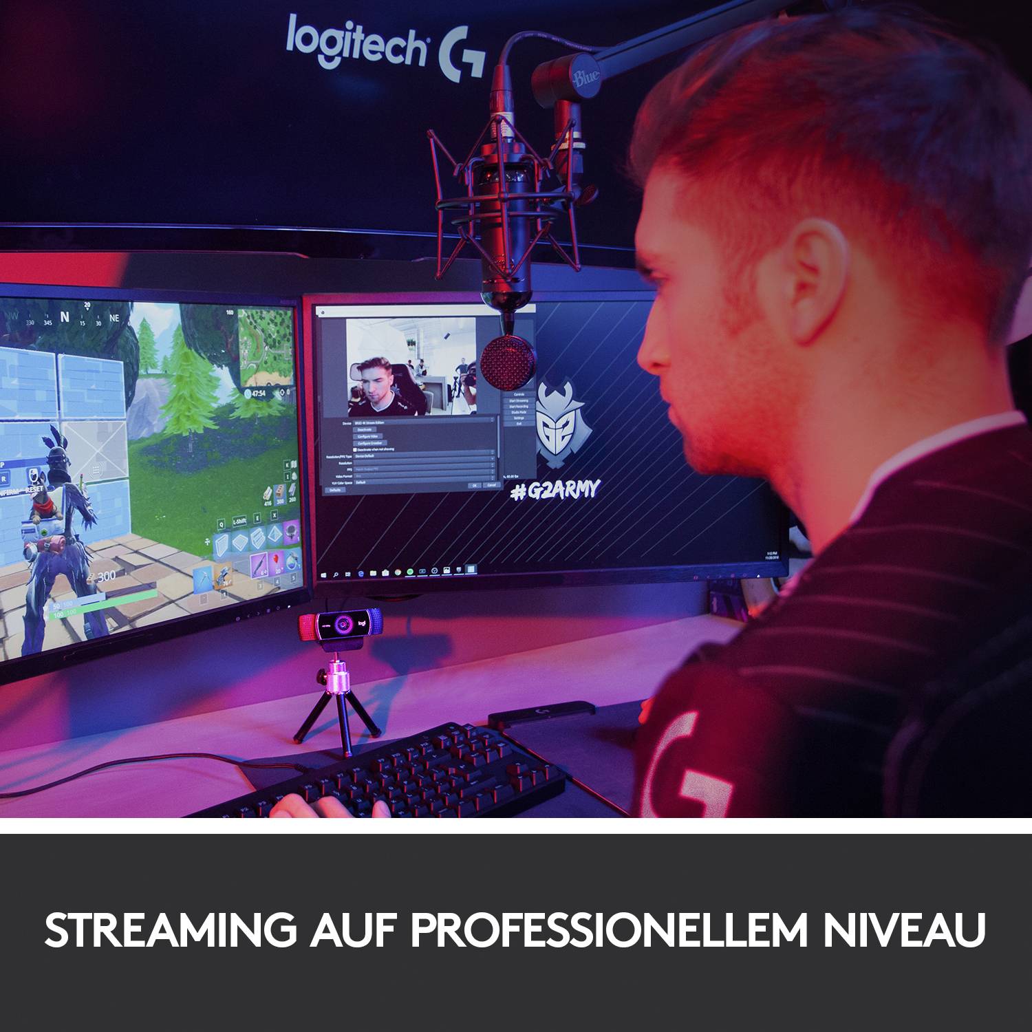 Ein Mann sitzt an einem Schreibtisch und streamt ein Videospiel auf zwei Monitoren. Oben ist ein Mikrofon; die Szene wirkt professionell.
