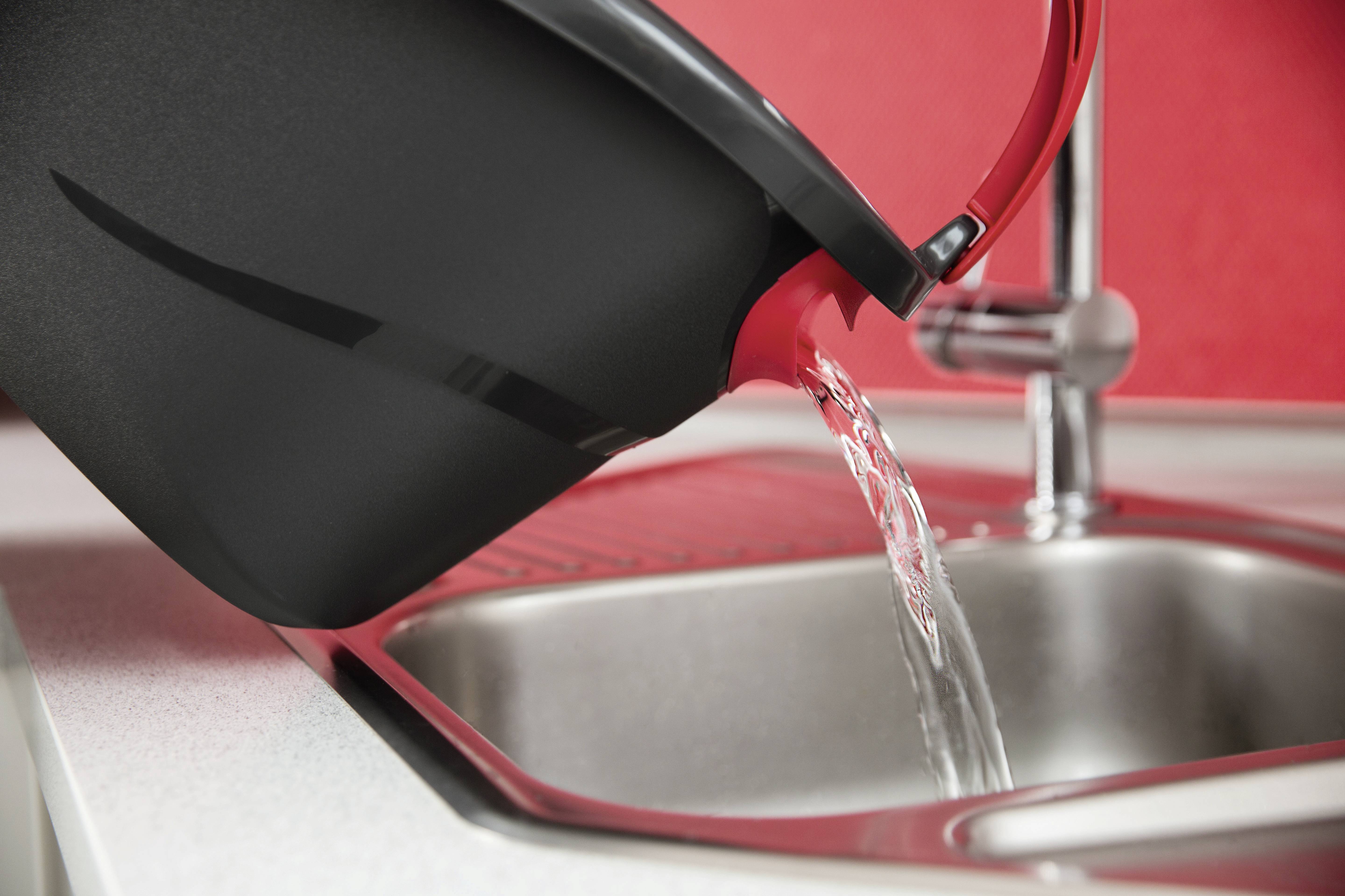 Eine schwarze Kanne mit rotem Griff gießt Wasser in ein silbernes Spülbecken. Im Hintergrund ein roter Rückwandbereich.