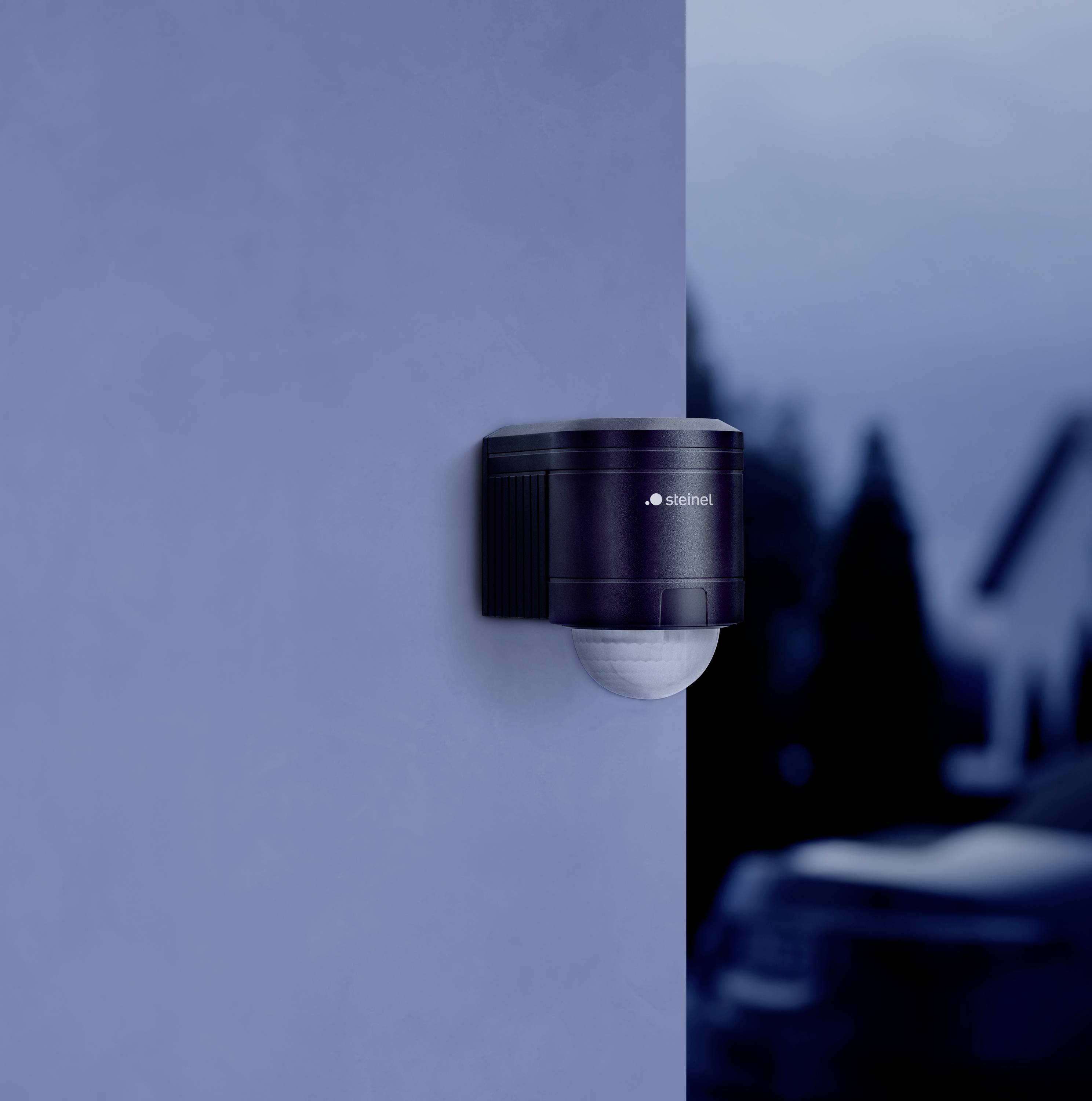 Eine schwarze Außenbewegungsmelder-Kamera an der Wand eines Gebäudes bei Dämmerung. Hintergrund zeigt unscharfen Gartenbereich.