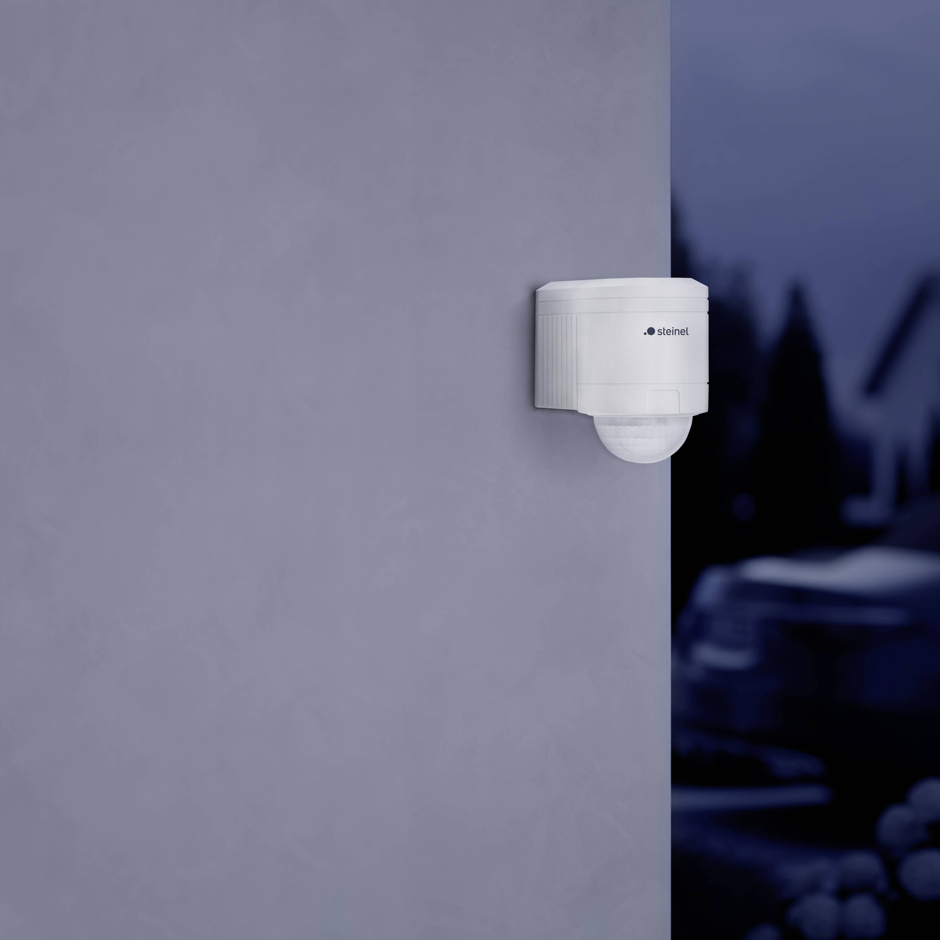 Ein Bewegungssensor ist an einer grauen Wand montiert. Im Hintergrund unscharf sichtbar ist ein Auto.