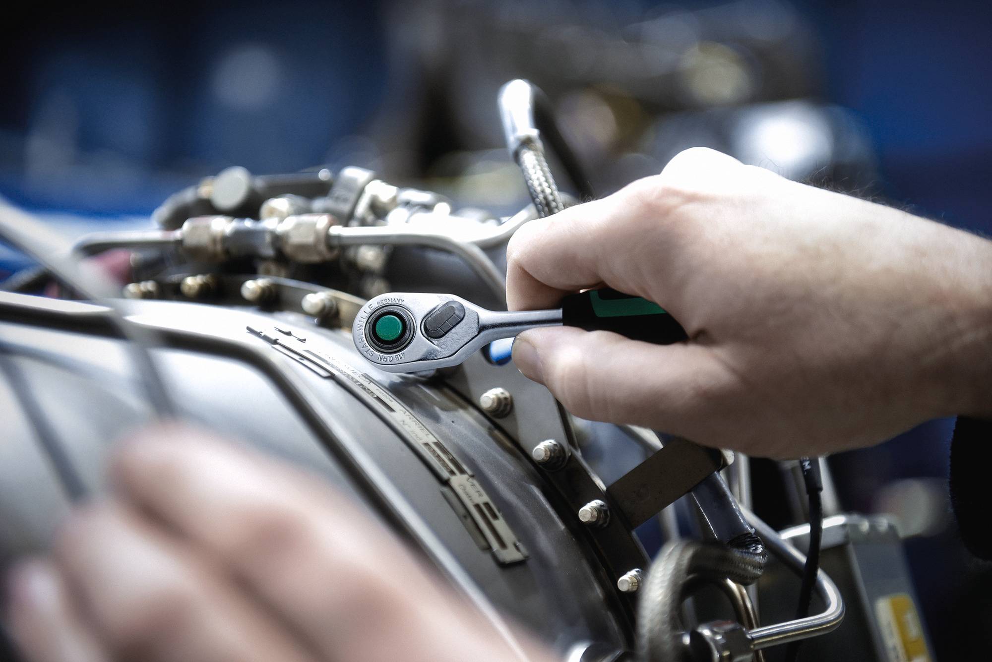 Eine Nahaufnahme einer Hand, die mit einem Schraubenschlüssel einen Bolzen an einem mechanischen Motor festzieht, umgeben von verschiedenen Rohren und Kabeln.