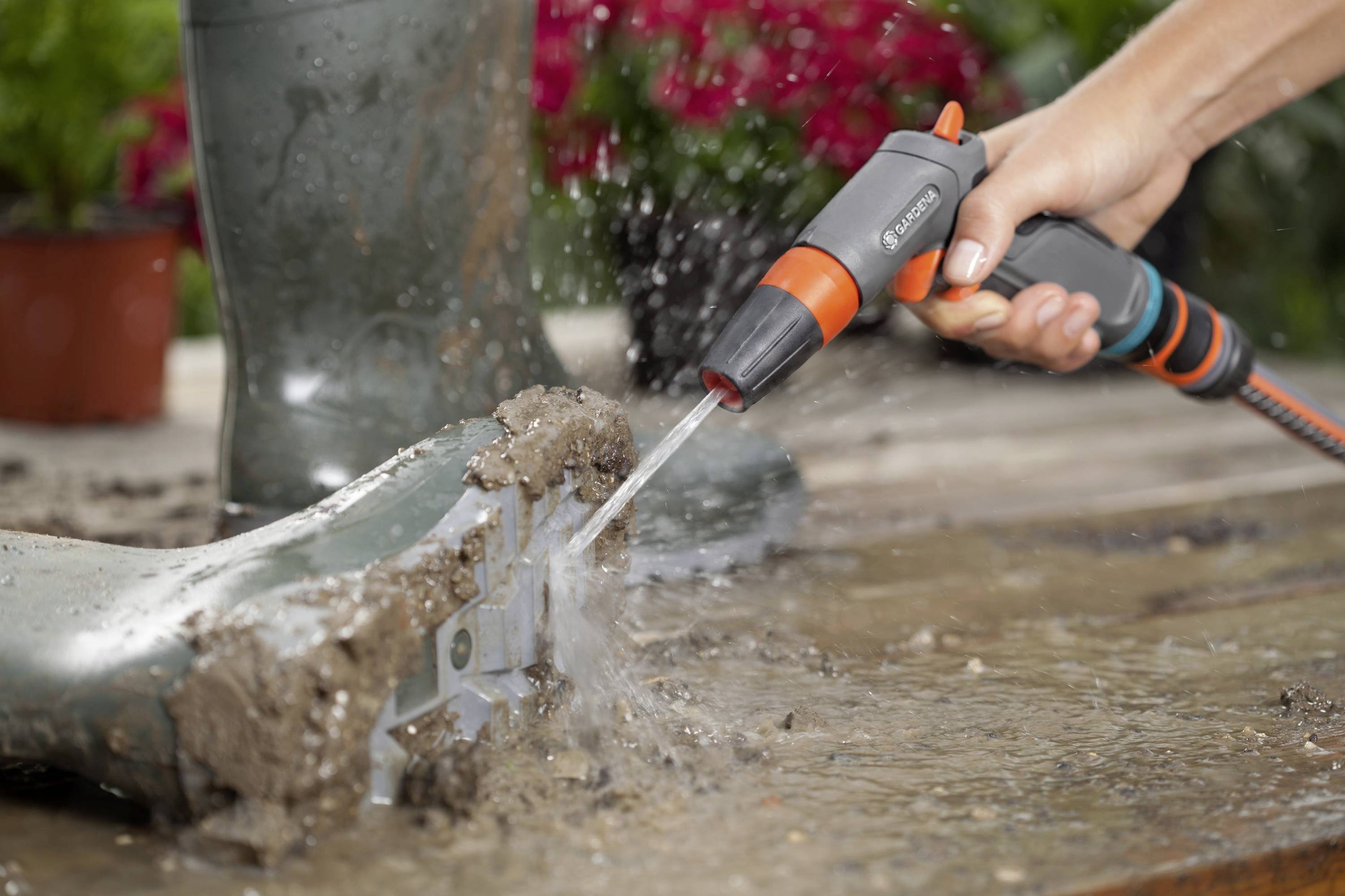 Ein Gartenschlauch spritzt Wasser auf einen schmutzigen Stiefel, um ihn zu reinigen. Im Hintergrund sind Pflanzen unscharf zu sehen.