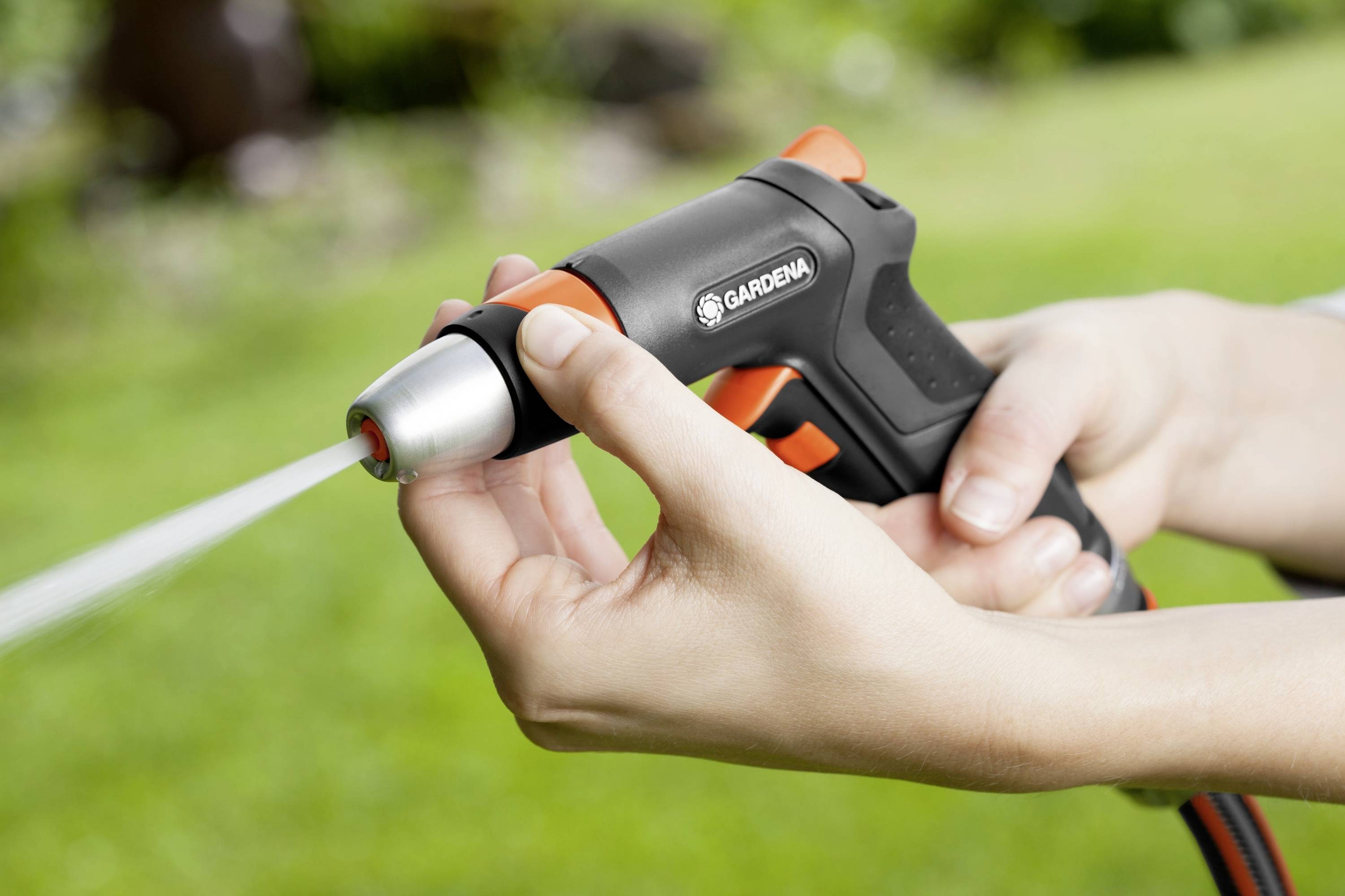 Person hält eine schwarze und orangefarbene Gartenschlauchdüse von Gardena, Wasser sprüht heraus, Hintergrund mit unscharfem Gras.