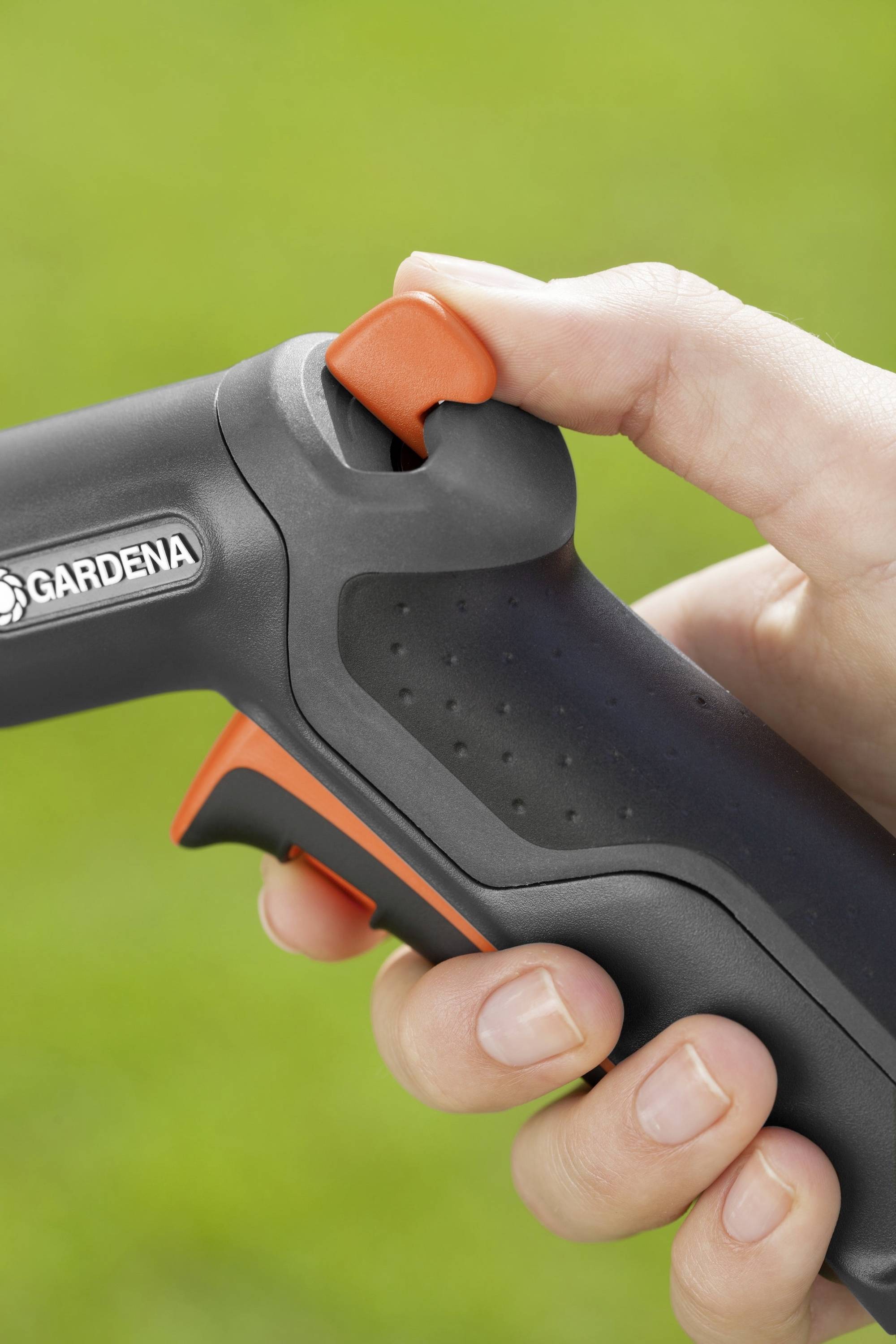 Person hält eine schwarze Gartenschlauchdüse von Gardena mit orangem Hebel, bereit zum Sprühen, auf grünem Hintergrund.