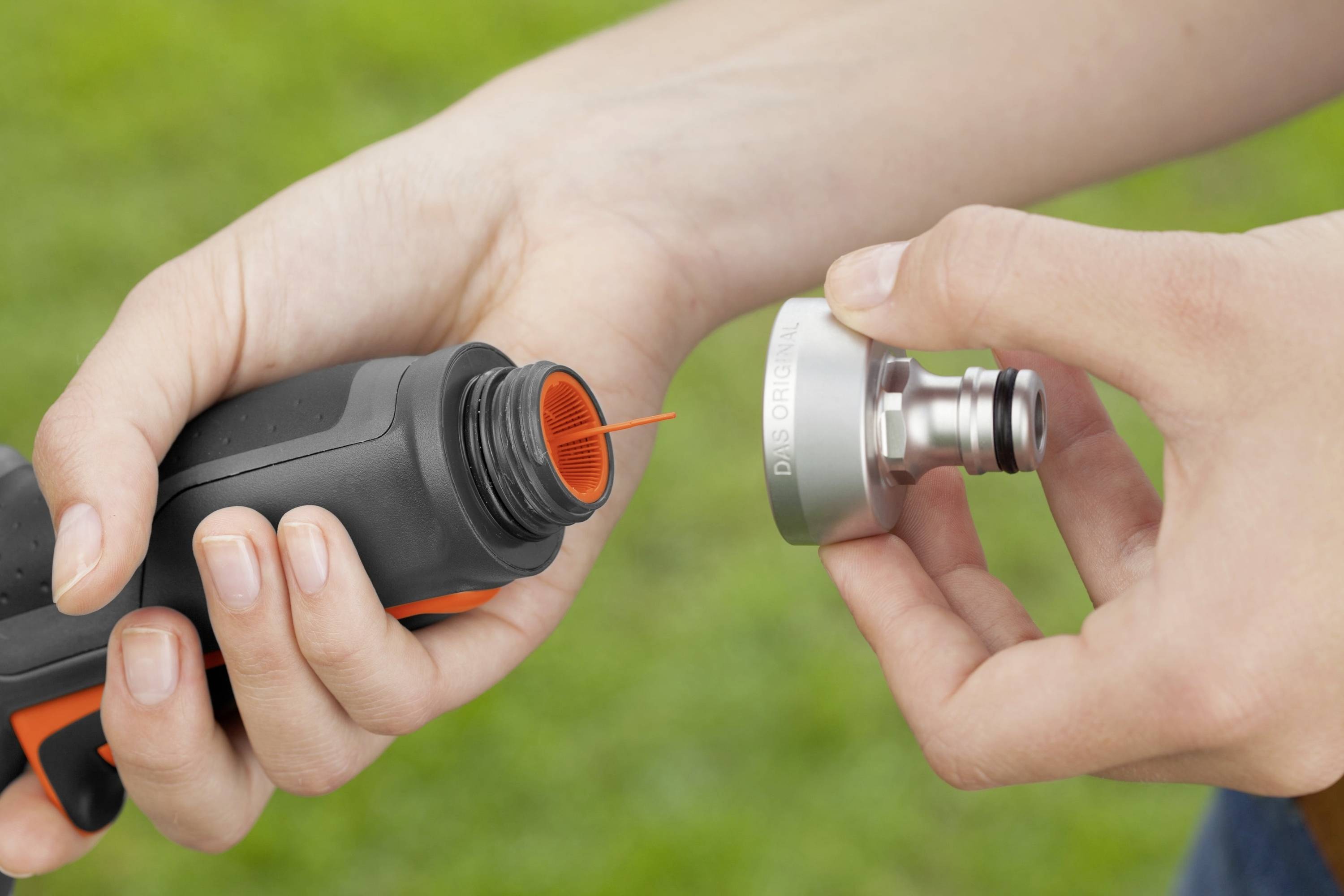 Eine Person hält Ausrüstungsteile in den Händen. Links ist ein schwarzes, flaschenähnliches Objekt mit oranger Düse, rechts ein metallisches Ventil.