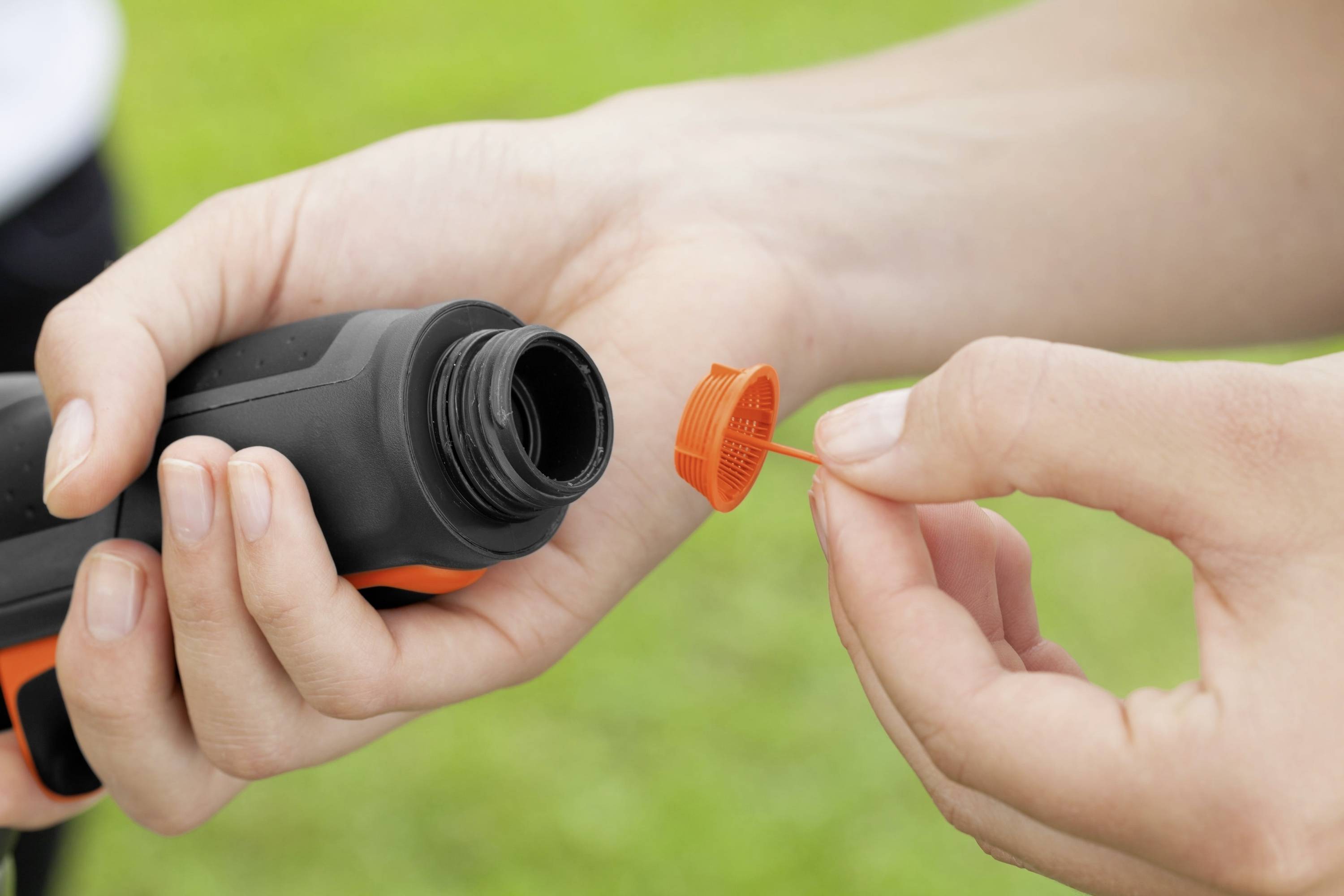 Person hält eine Flasche mit abgenommenem orangefarbenem Deckel im Grünen.