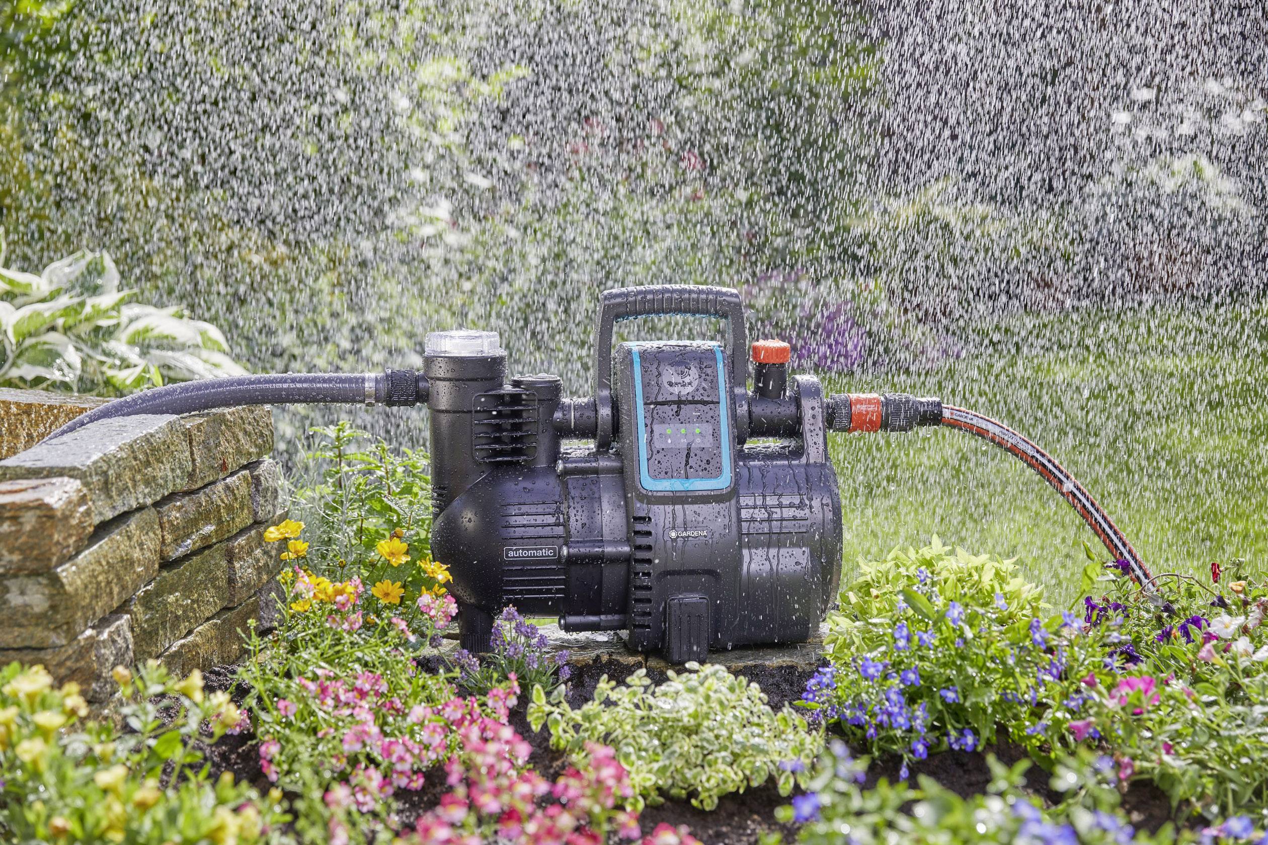 Eine Bewässerungspumpe versorgt einen blühenden Garten mit Wasser. Regenähnliche Gischt verteilt sich über Blumen in verschiedenen Farben.