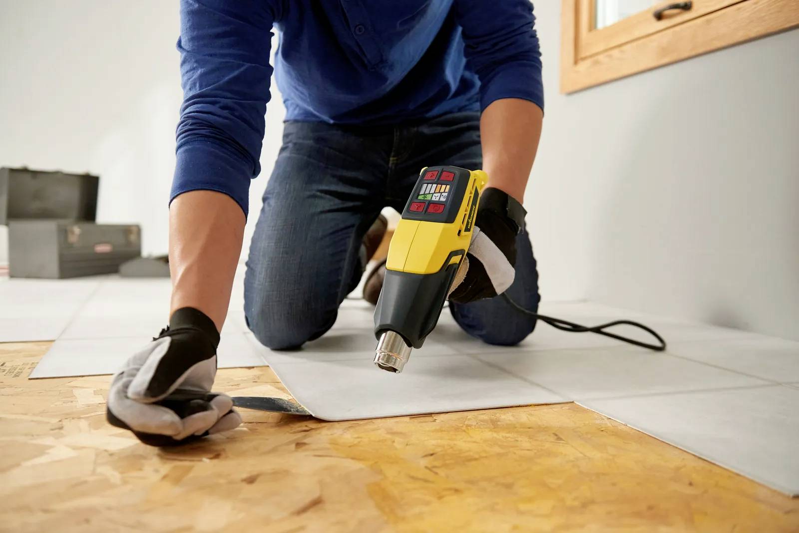 Eine Person kniet auf dem Boden und verwendet eine Heißluftpistole, um Bodenbeläge zu installieren. Sie trägt Handschuhe zum Schutz.