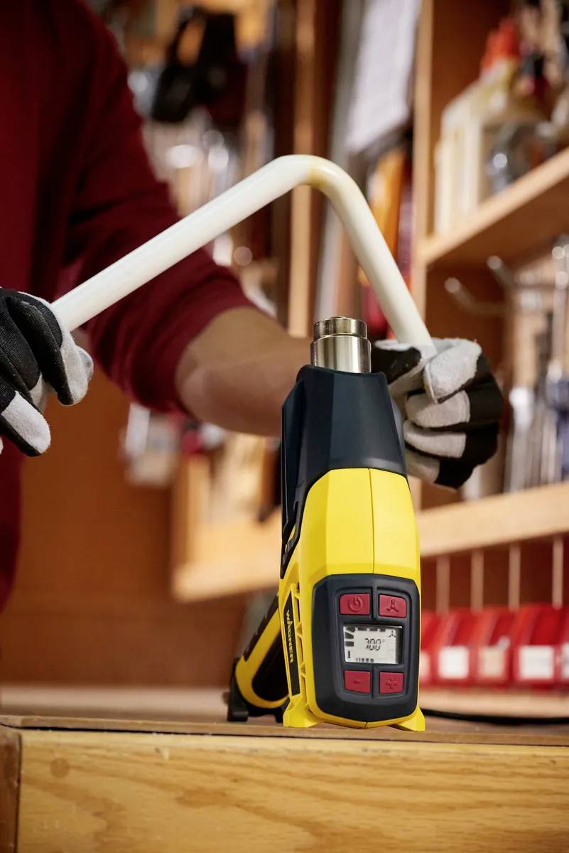 Eine Person biegt mit einem gelben Heißluftgerät ein weißes Rohr in einer Werkstatt. Das Gerät zeigt eine Temperatur von 300°C an.
