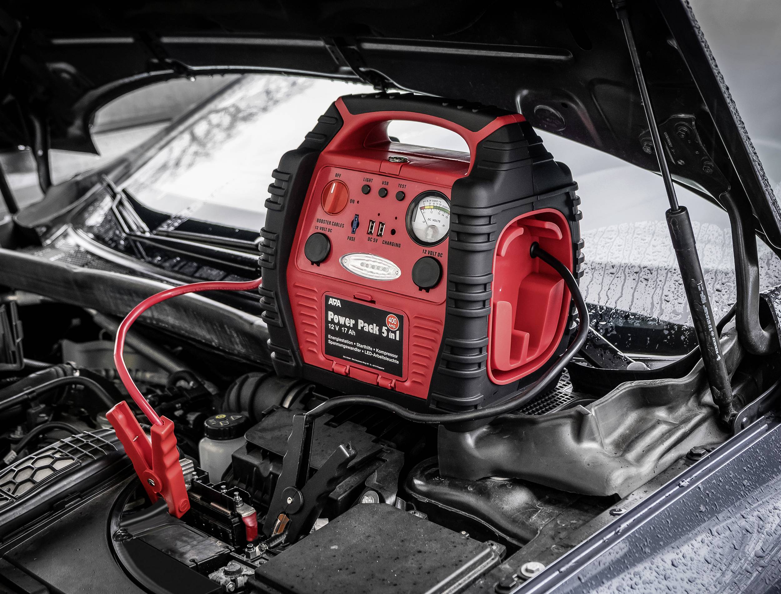 Starthilfe-Booster auf einer Autobatterie unter geöffneter Motorhaube. Rote und schwarze Kabel mit Klemmen sind angeschlossen.