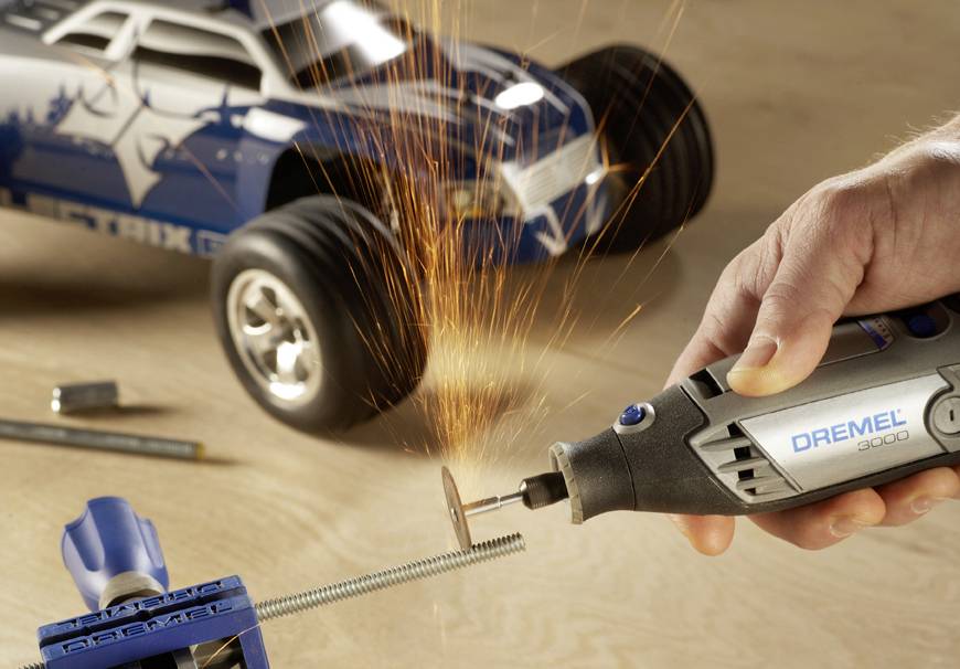 Eine Hand hält ein Dremel-Werkzeug, das Funken sprüht, während es eine Schraube schneidet. Im Hintergrund steht ein Modellauto.