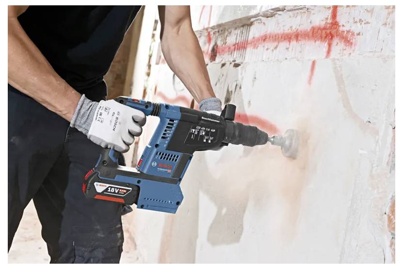 Eine Person, die eine blaue Bohrmaschine mit einem schwarzen Griff an einer Betonwand verwendet. Rote Markierungen und Staub sind an der Wand zu sehen.