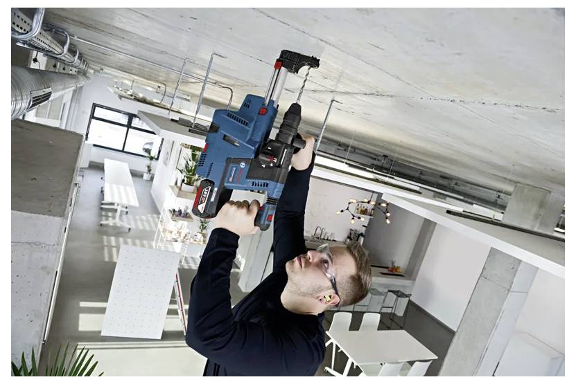 Eine Person benutzt einen elektrischen Bohrer, um an einer Decke in einem industriell gestalteten Raum mit Tischen und Stühlen zu arbeiten, und trägt dabei Schutzbrille und Ohropax.