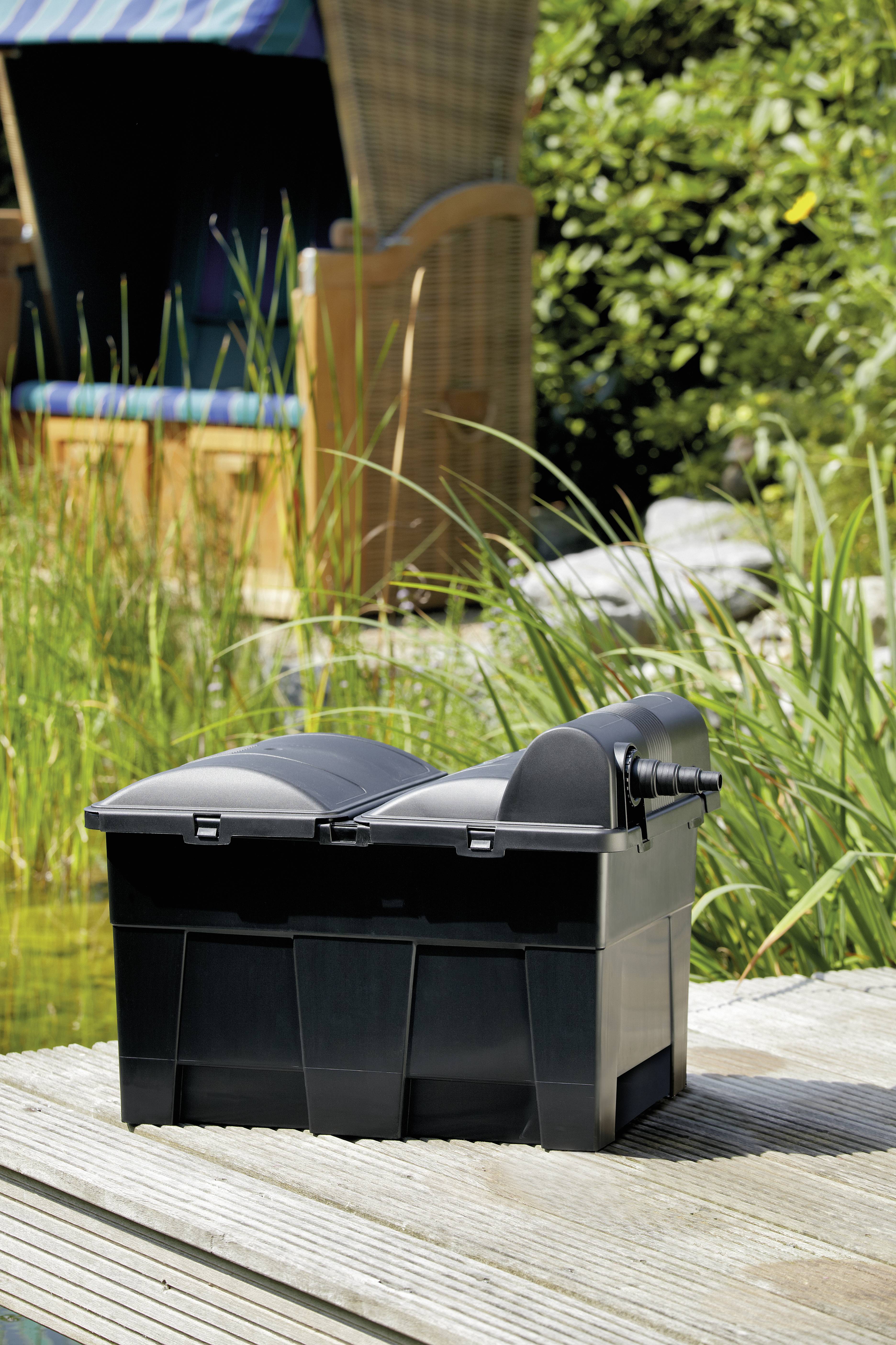 Eine schwarze Filterbox, die auf einer Holzterrasse neben einem Teich platziert ist, mit hohem Gras und einem überdachten Stuhl im Hintergrund im Freien.