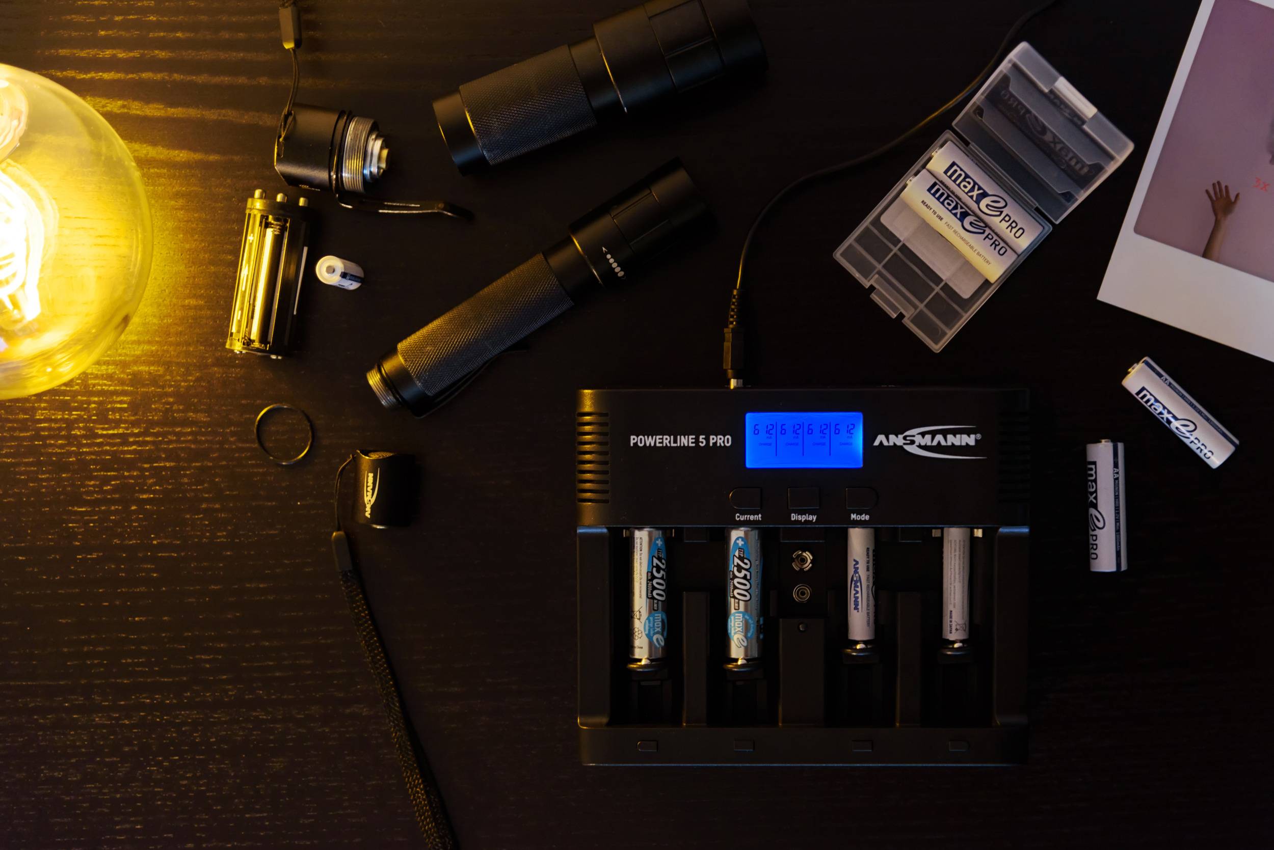 Ein Batterie-Ladegerät lädt mehrere AA-Batterien auf einem Tisch. Daneben liegen Taschenlampen, ein Akku-Behälter und ein Foto.