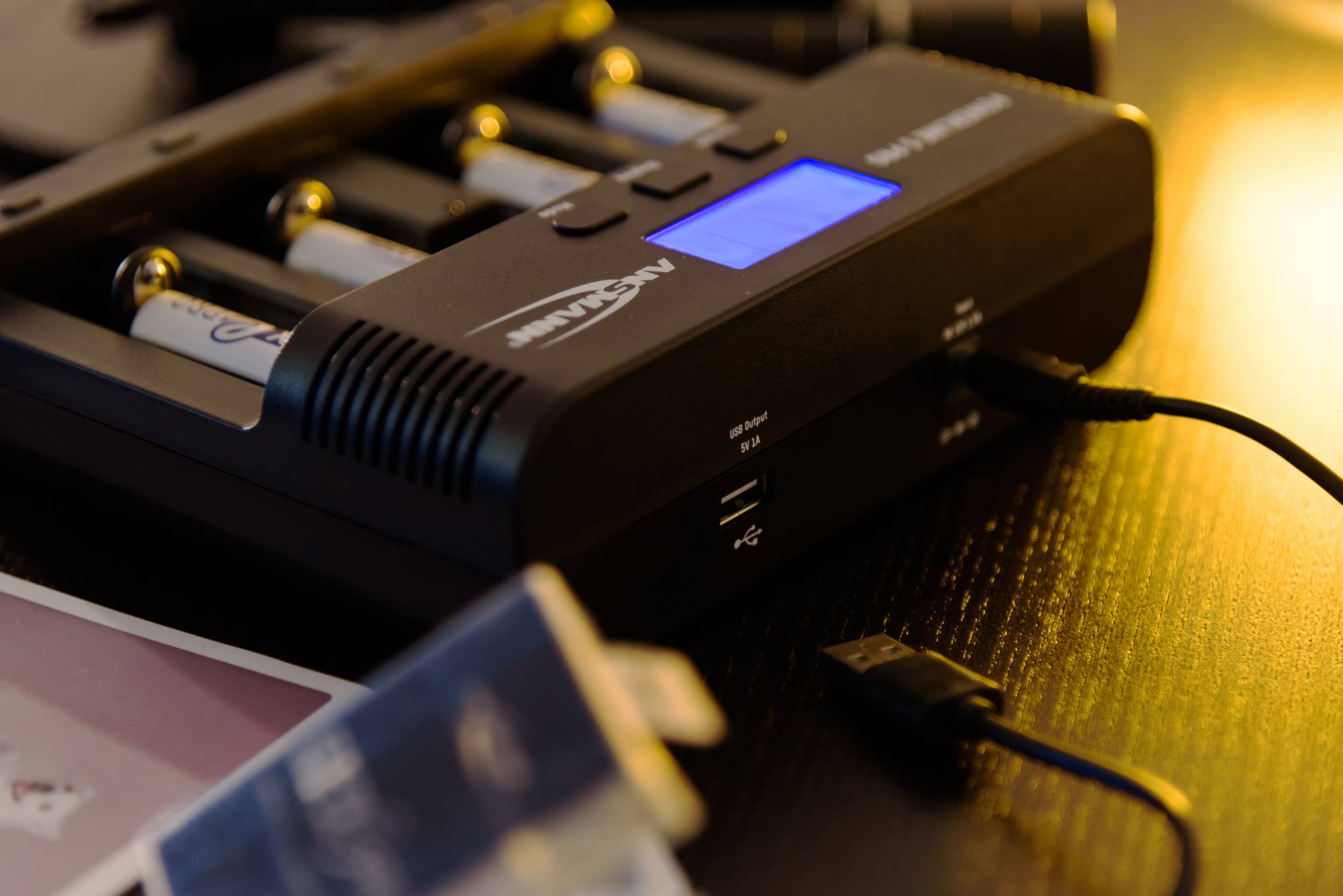 Ladegerät mit vier eingelegten AA-Batterien und blauem Display, zeigt Ladezustand an, umgeben von USB-Kabel und unscharfem Hintergrund.