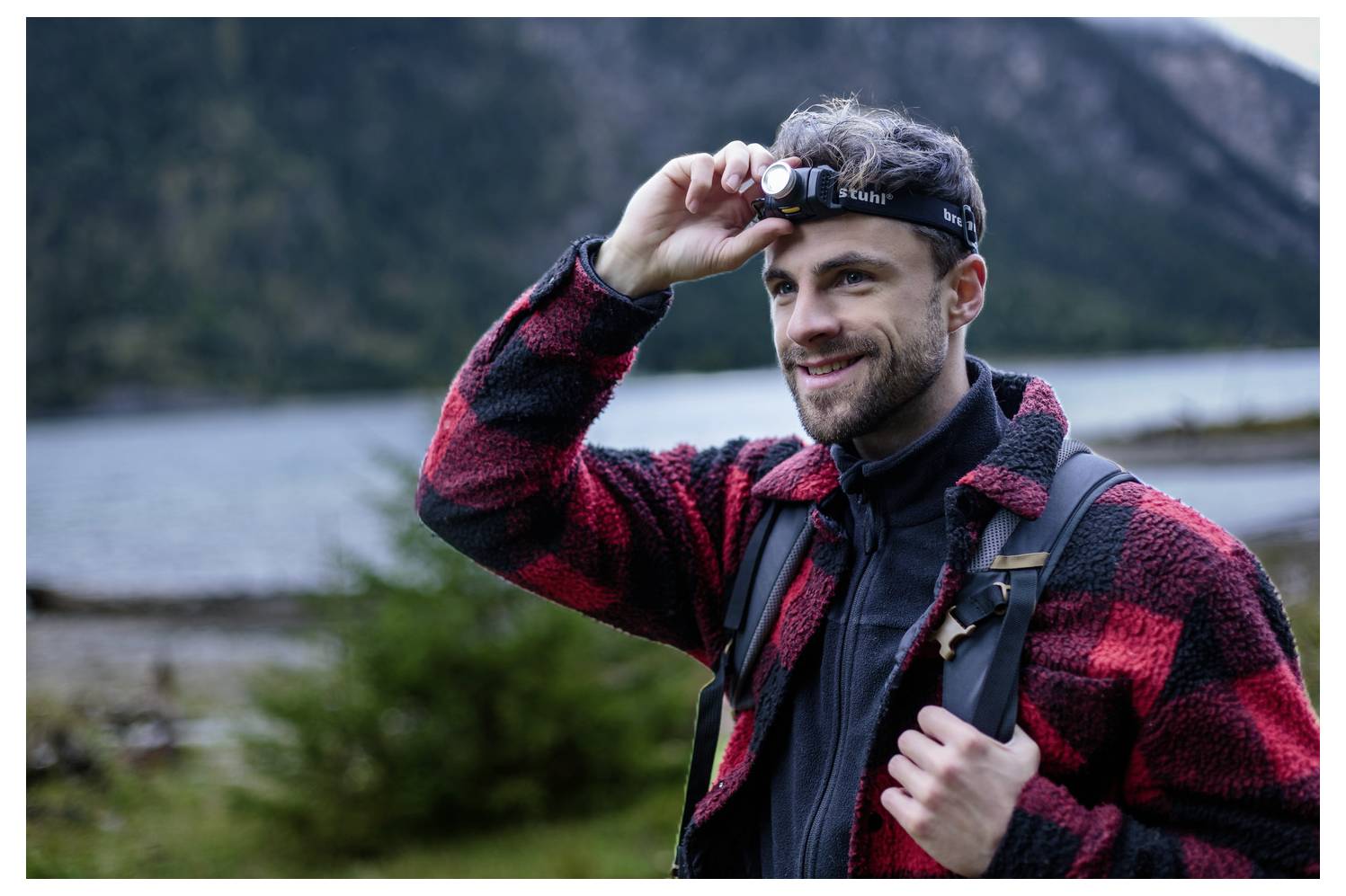 Ein lächelnder Mann im Freien an einem See stellt eine Stirnlampe auf seiner Stirn ein, während er eine rote Karohemd und einen Rucksack trägt.