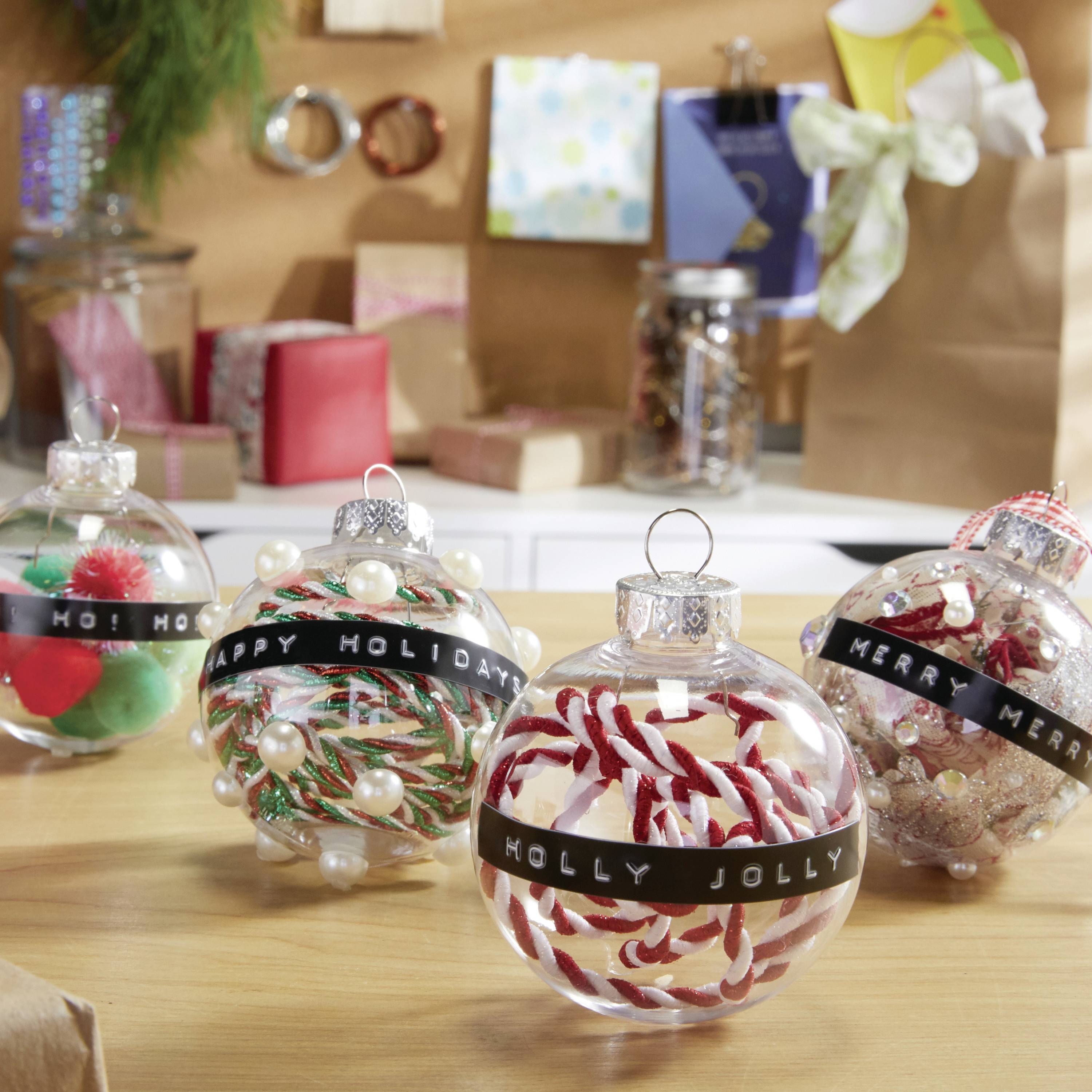 Vier durchsichtige Weihnachtskugeln auf einem Tisch, gefüllt mit verschiedenen Dekorationen wie Pompons und Streifen, mit Feiertagsgrüßen.