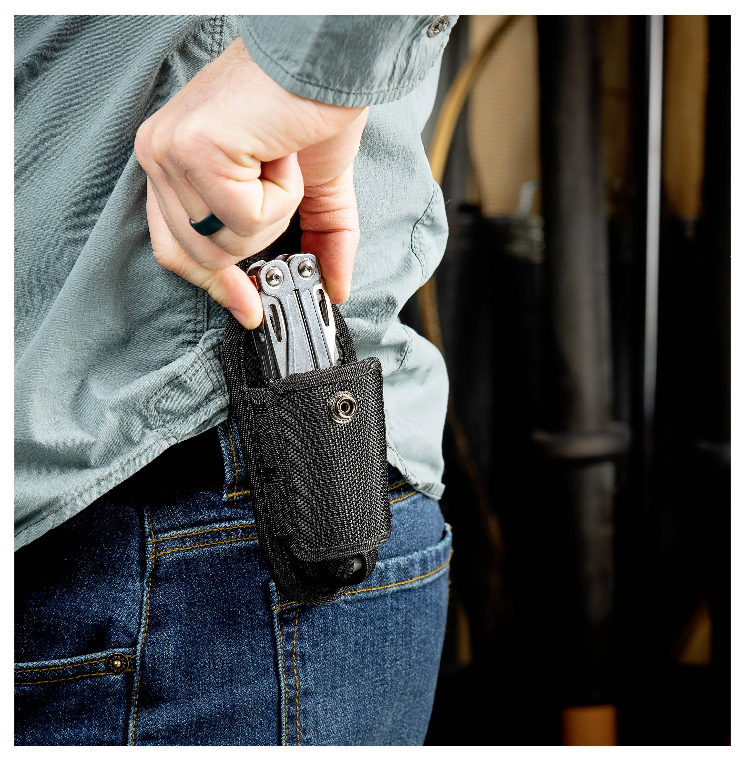 Person mit blauen Jeans und einem grünen Hemd, die ein Multitool mit einer schwarzen Tasche an der Seite hält.