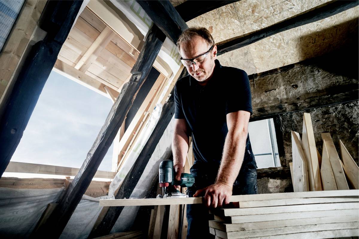 Ein Mann trägt Schutzbrille und bohrt in einem Dachboden Holzplanken. Sonnenlicht fällt durch ein teilweise geöffnetes Fenster.