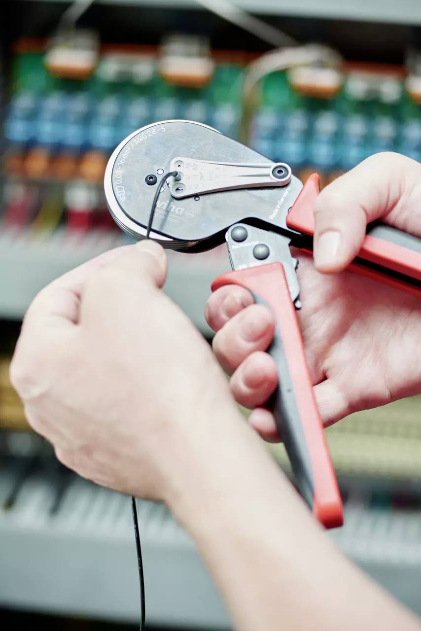 Eine Person benutzt eine Crimpzange, um ein Kabel zu bearbeiten, vor einem unscharfen elektrischen Schaltschrank im Hintergrund.