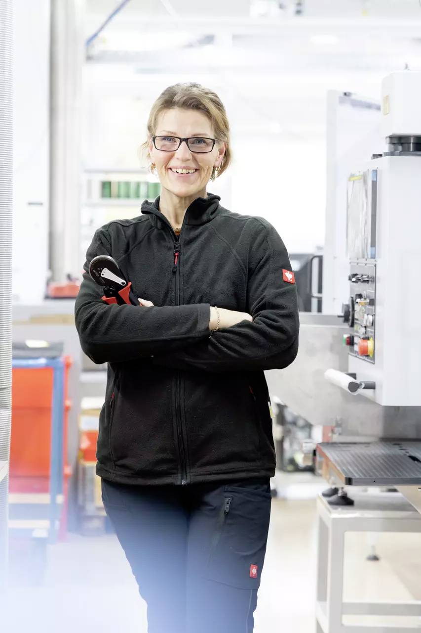 Eine Frau in Arbeitskleidung steht in einer Werkstatt, hält ein Werkzeug und lächelt in die Kamera. Im Hintergrund sind Maschinen erkennbar.