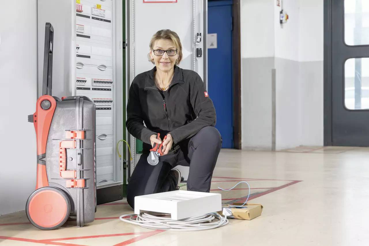 Eine Frau kniet vor einem Schaltschrank und hält ein Messgerät in der Hand. Neben ihr steht ein roter Koffer mit Rollen.