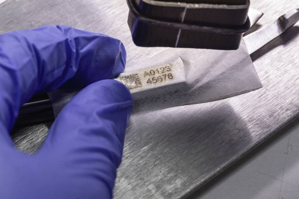 'Eine Person hält eine Folie unter einem Werkzeug auf einem Metalltisch. Auf der Folie stehen die Nummern A0123 und 45678.'