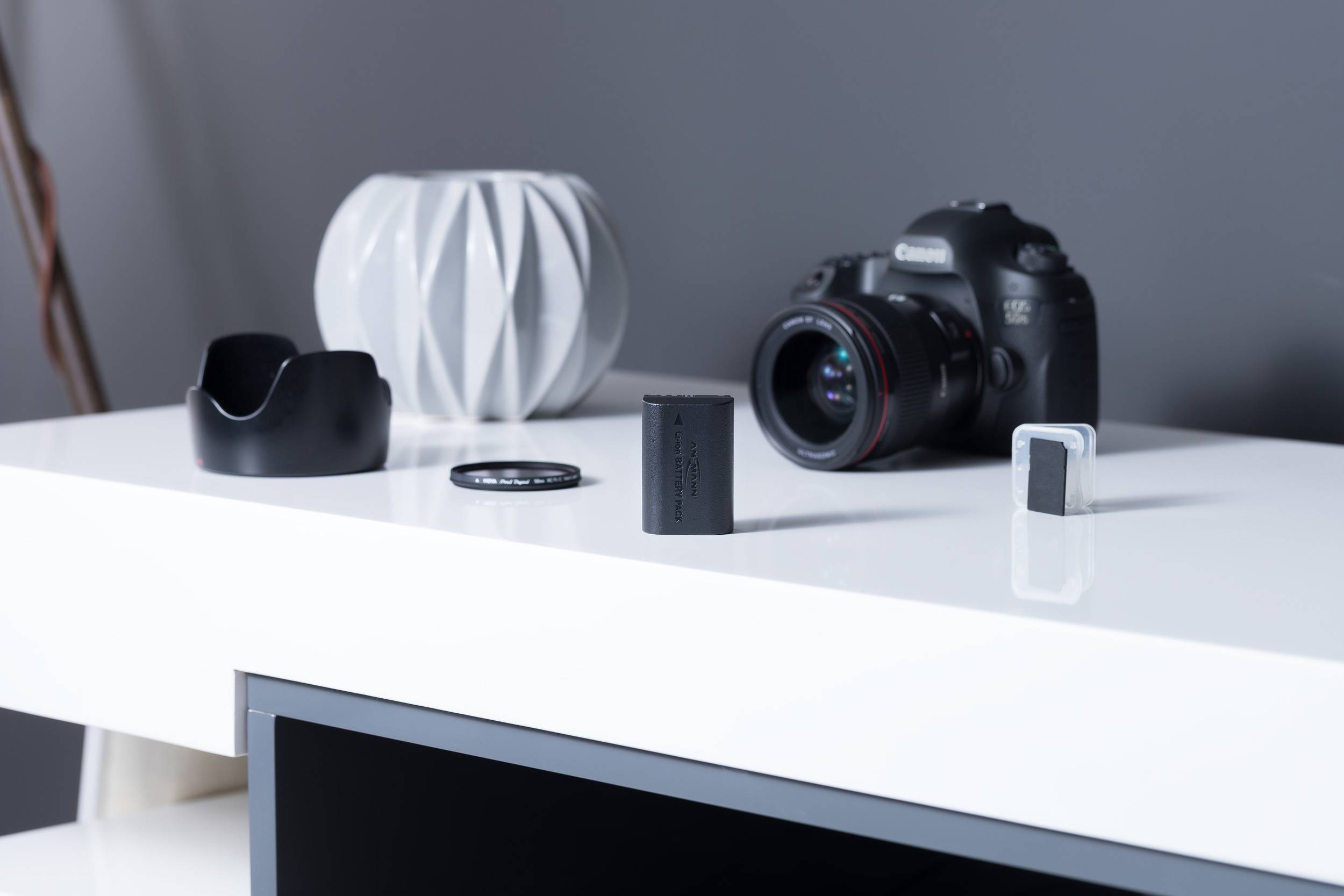 Eine Kamera mit Objektiv sitzt auf einem weißen Tisch. Daneben liegen eine Linsenabdeckung, ein Batteriepack und ein grauer Dekorartikel.