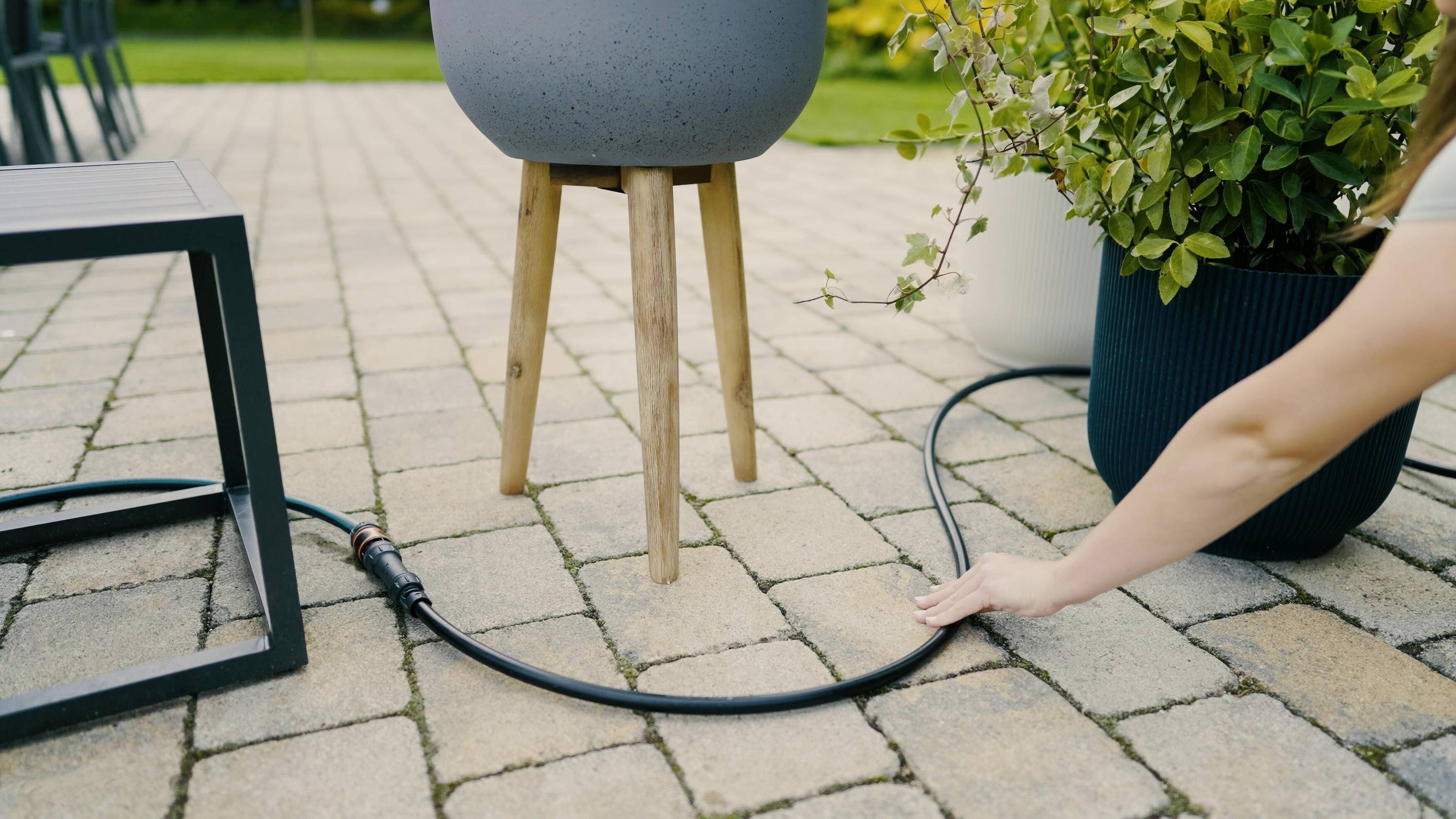 Eine Person richtet einen Gartenschlauch auf einer gepflasterten Terrasse aus, neben zwei Pflanzgefäßen und einem Tisch im Freien.