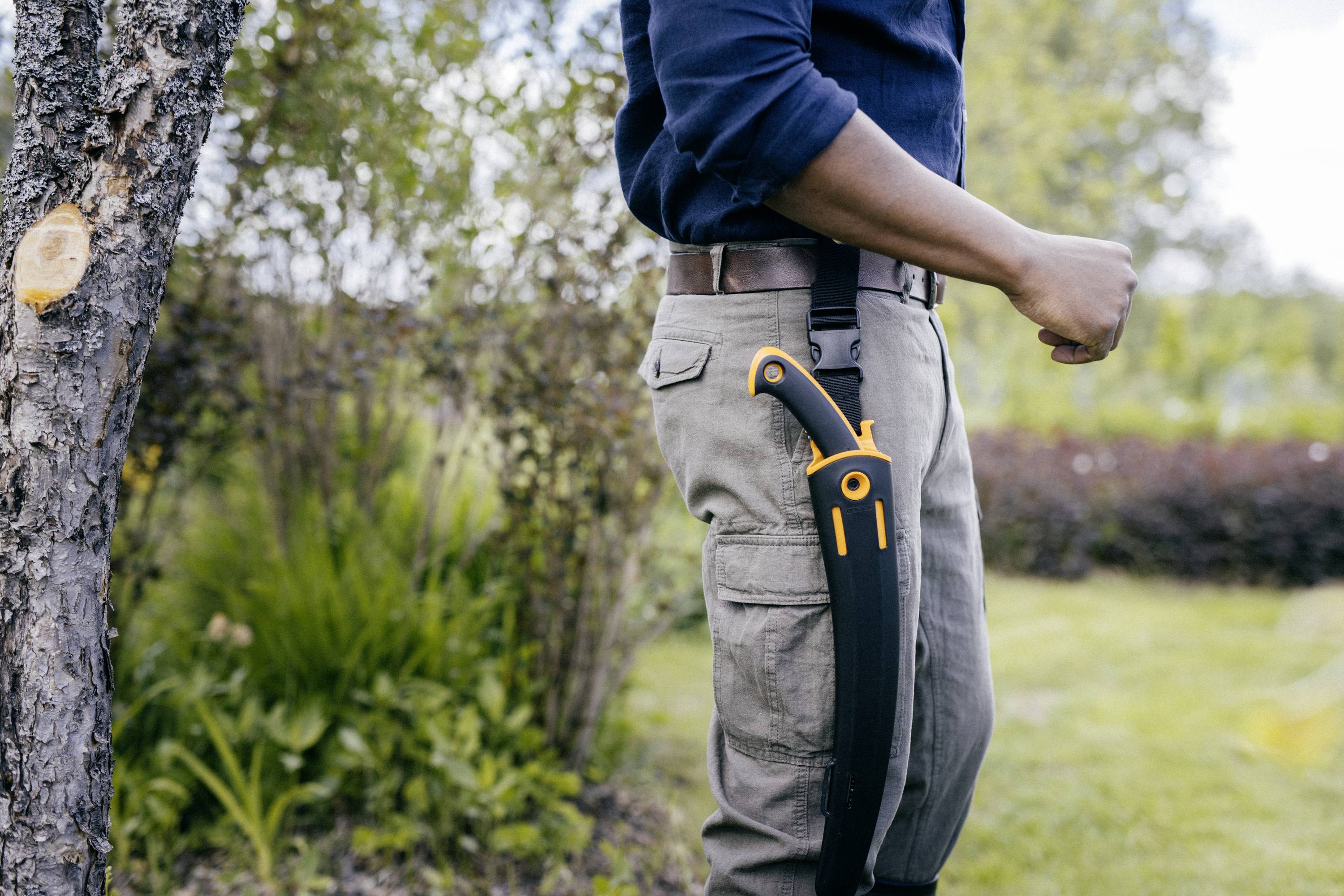 Mann steht im Garten, trägt Messer an der Seite, bereit für Gartenarbeit. Hintergrund zeigt Bäume und Rasen.