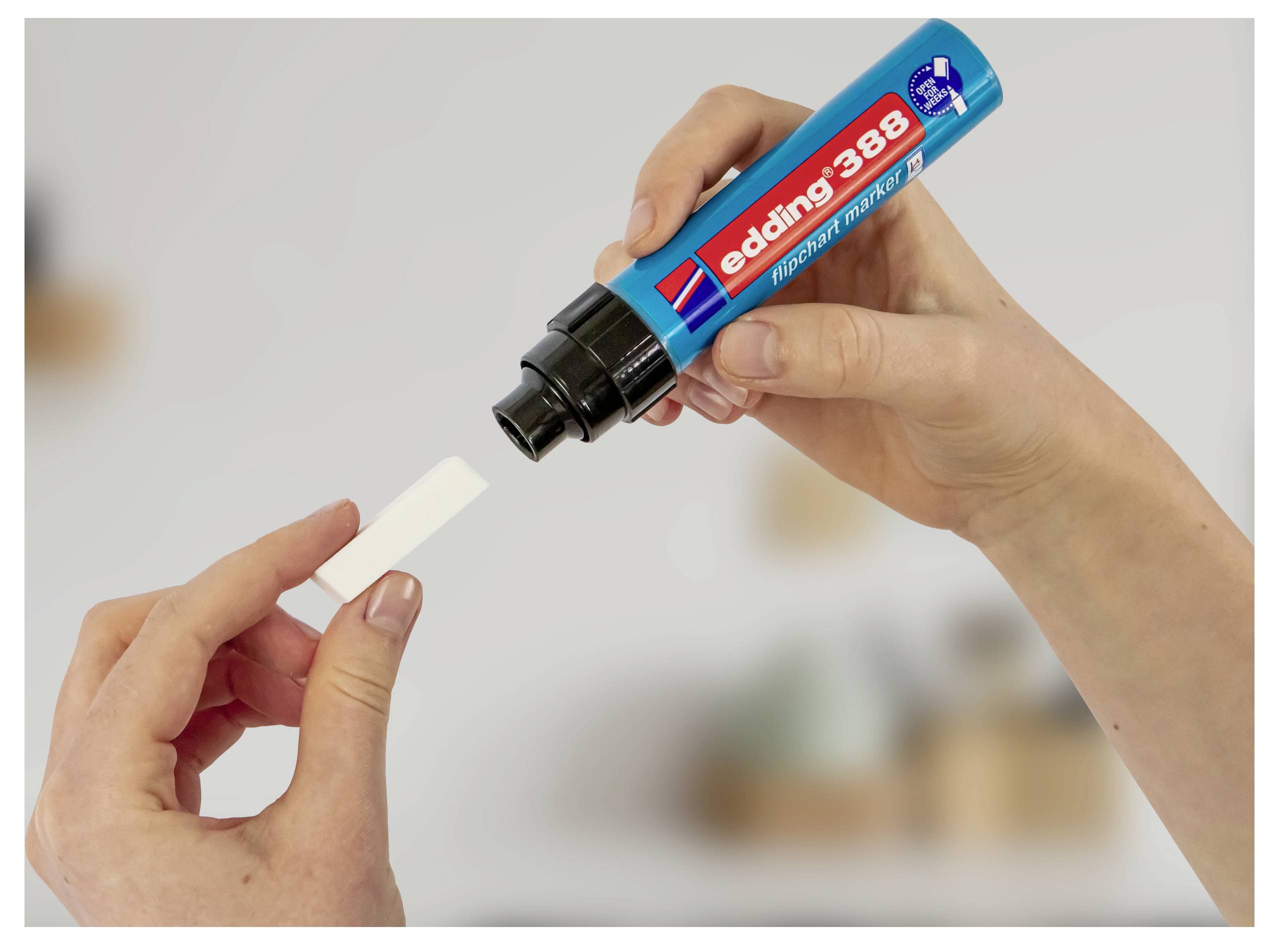 Hände, die mit einem blauen „Edding 388"-Marker auf ein kleines weißes Etikett schreiben, in Nahaufnahme vor unscharfem Hintergrund gezeigt.