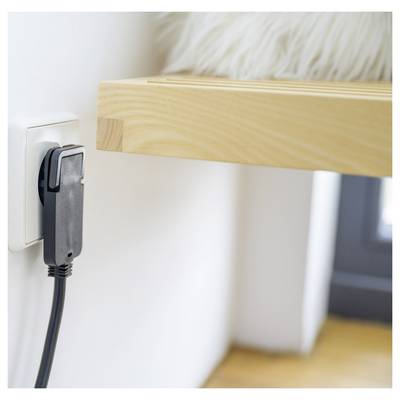 Nahaufnahme eines schwarzen Stromsteckers, der in eine Wandsteckdose neben einem Holzregal eingesteckt ist, mit Kabel, das nach unten hängt.