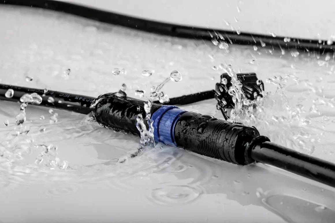 Ein wasserdichtes Kabel mit einem blauen Verbindungsstück liegt auf einer nassen Oberfläche, Wasser spritzt darauf.