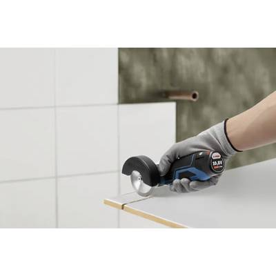 Eine Person mit Handschuhen verwendet eine elektrische Handkreissäge, um die Kante einer weißen Kachel zu schneiden, vor einem Hintergrund von gefliesten und strukturierten Wänden.
