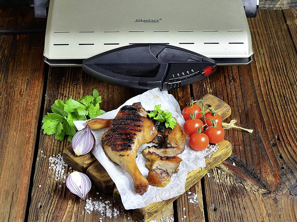 Gegrilltes Hähnchenbein mit Tomaten, Zwiebel und Petersilie auf einem Holzbrett vor einem Tischgrill auf einem Holztisch.