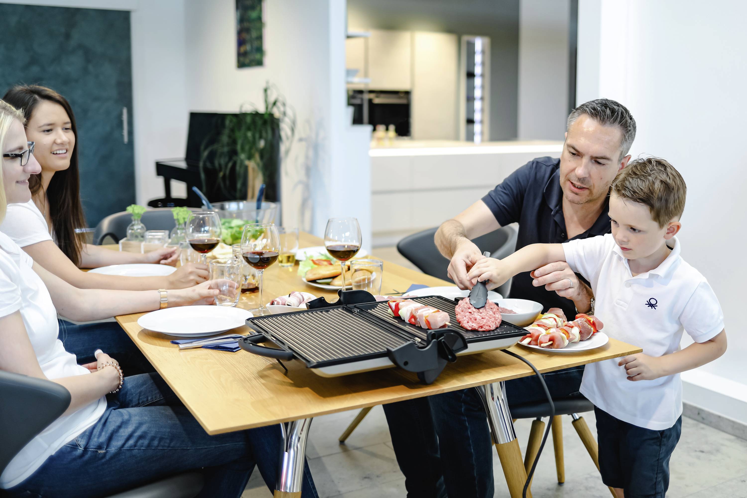 Eine Familie sitzt an einem Tisch und grillt zusammen drinnen. Ein Mann und ein Kind legen Fleisch auf einen Tischgrill, während andere zuschauen.