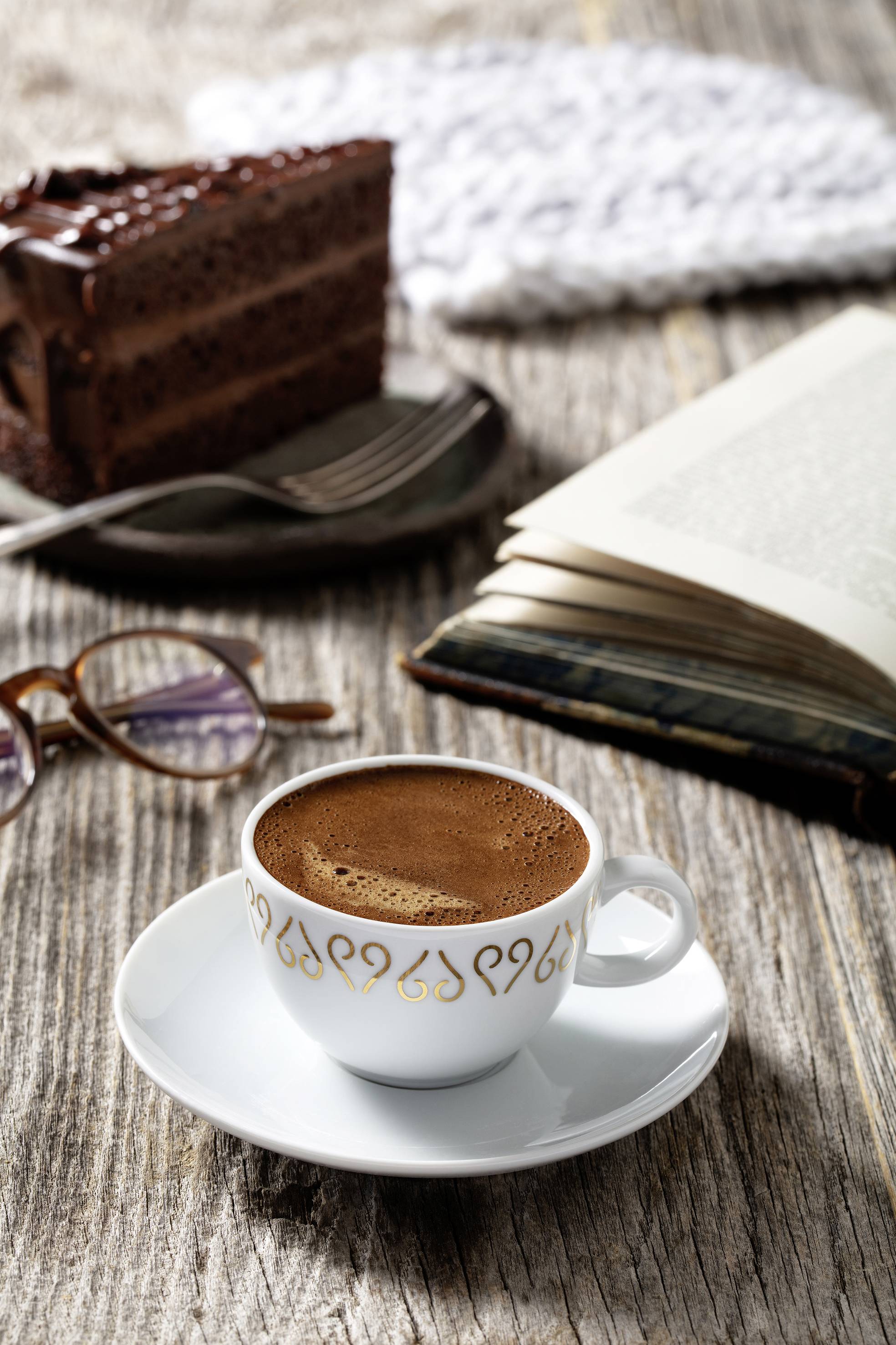 Tasse Kaffee auf Holztisch, daneben ein Buch, Brille und ein Stück Schokoladenkuchen. Die Szene wirkt gemütlich und einladend.