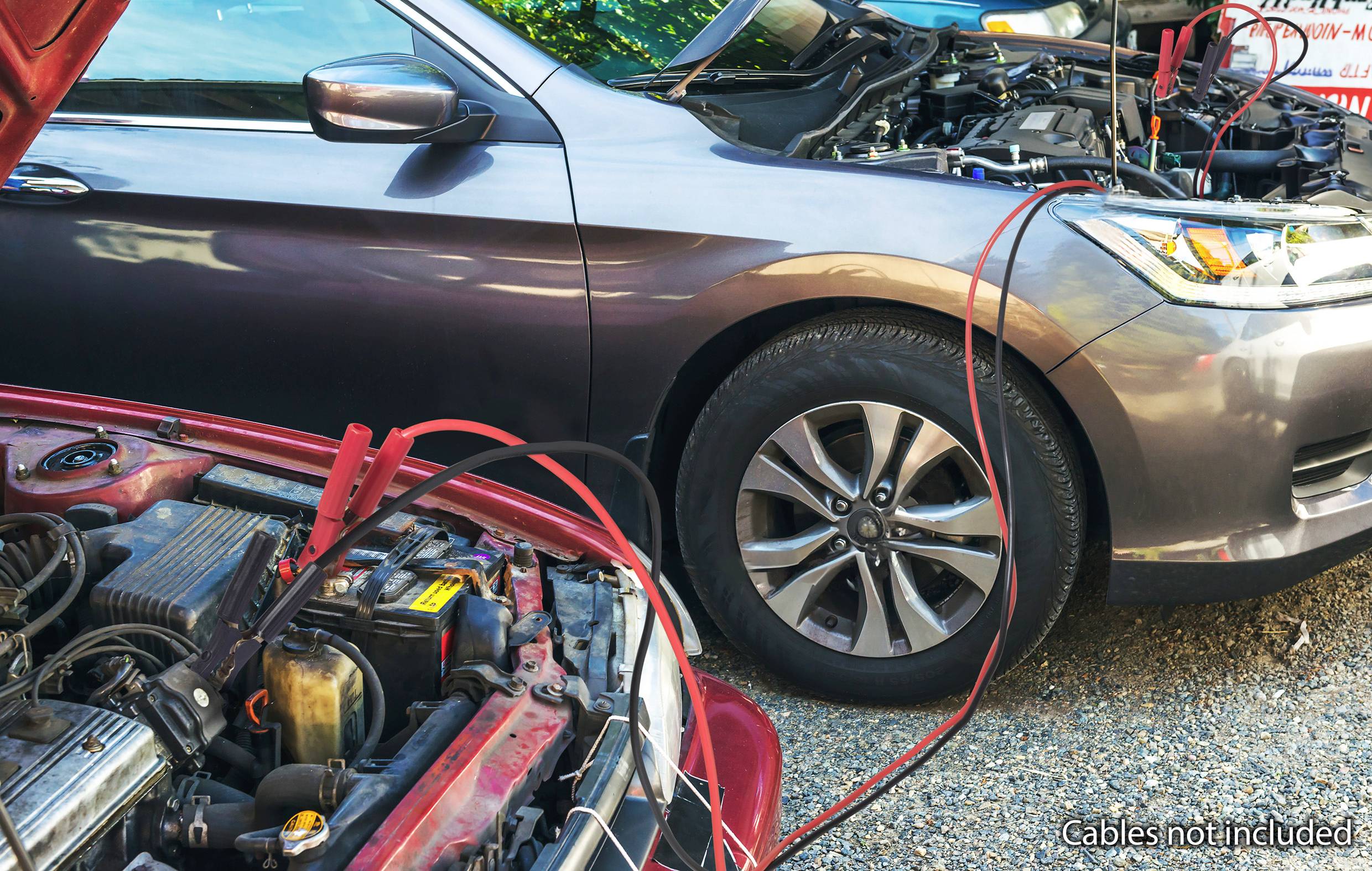 Zwei Autos mit geöffneten Motorhauben, verbunden durch rote Starthilfekabel, zeigen eine Starthilfesituation auf einem Kiesweg.